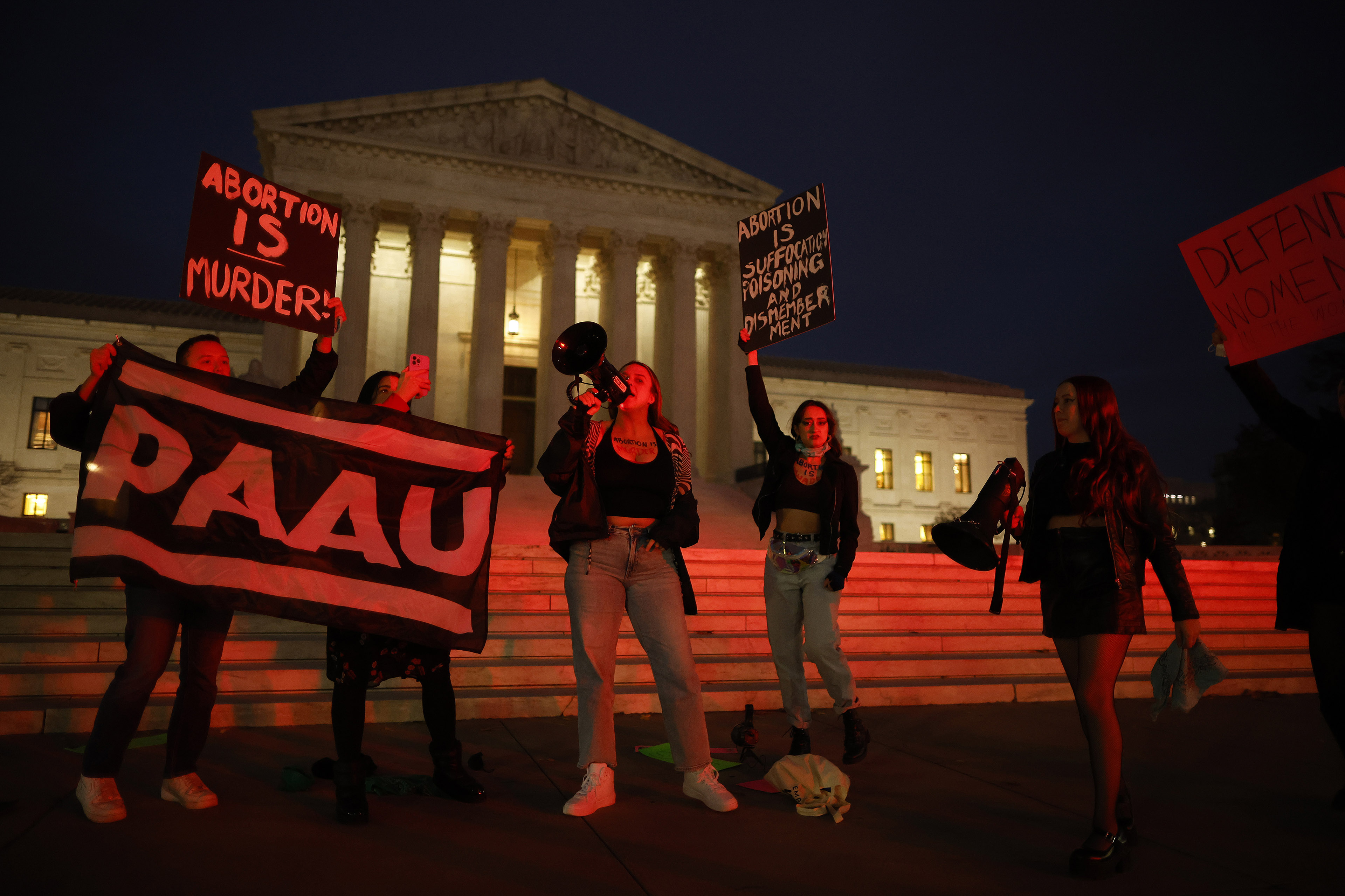 Members of the Progressive Anti-Abortion Uprising rally in front of the U.S. Supreme Court. They are illuminated by a strong red light. A woman standing in the center, while protesters around her hold signs with anti-abortion slogans.