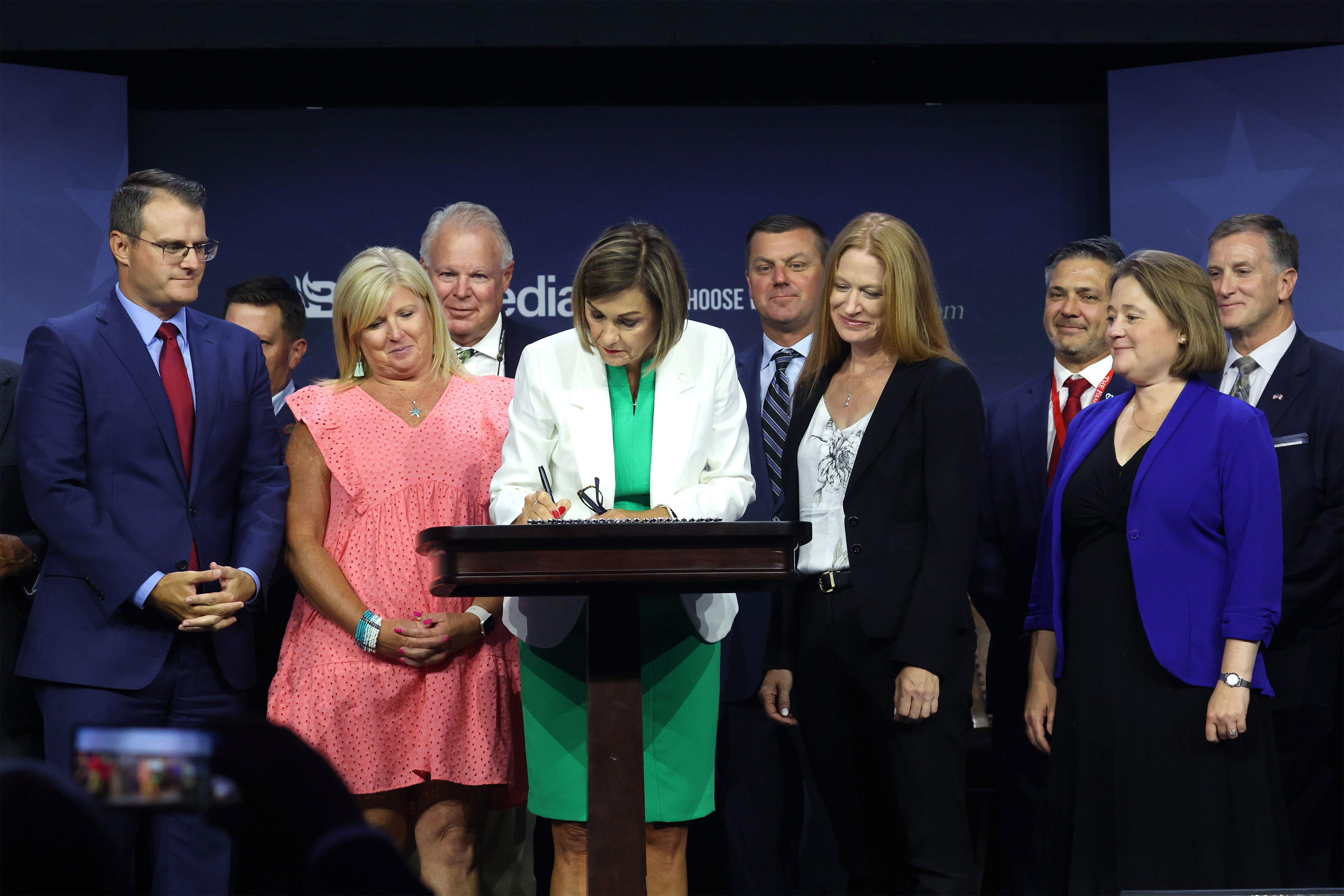 A photo of Iowa's governor, Kim Reynolds, signing a bill into law banning most abortions in the state after about six weeks. People are standing behind her and watching as she signs.