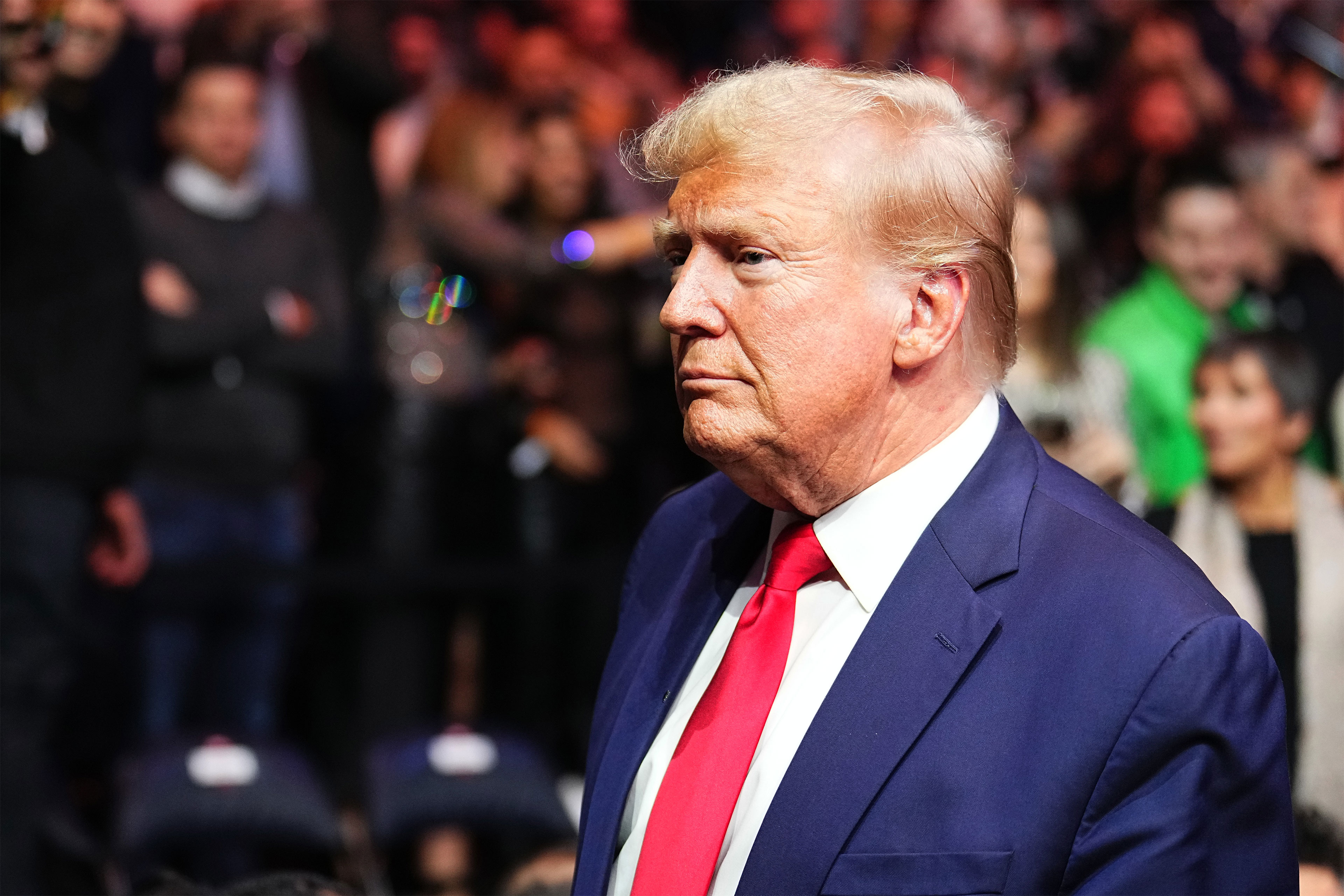 A man in a suit and red tie stands at an angle to the camera. There are many people behind him and out of focus.