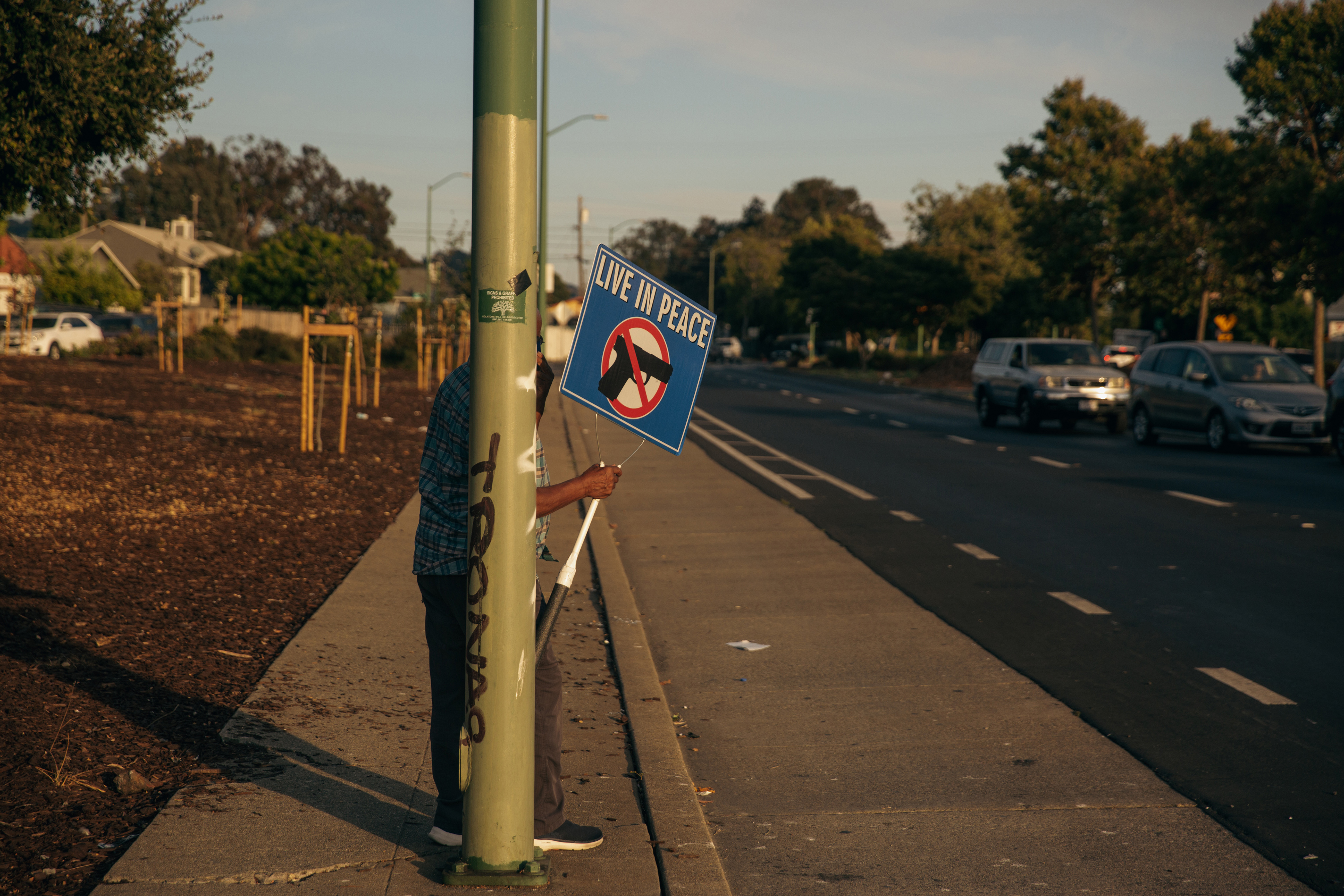 A person holds a sign that reads "Live In Piece" and has the image of a handgun behind a general prohibition sign. They stand behind a streetlight pole beside a road.