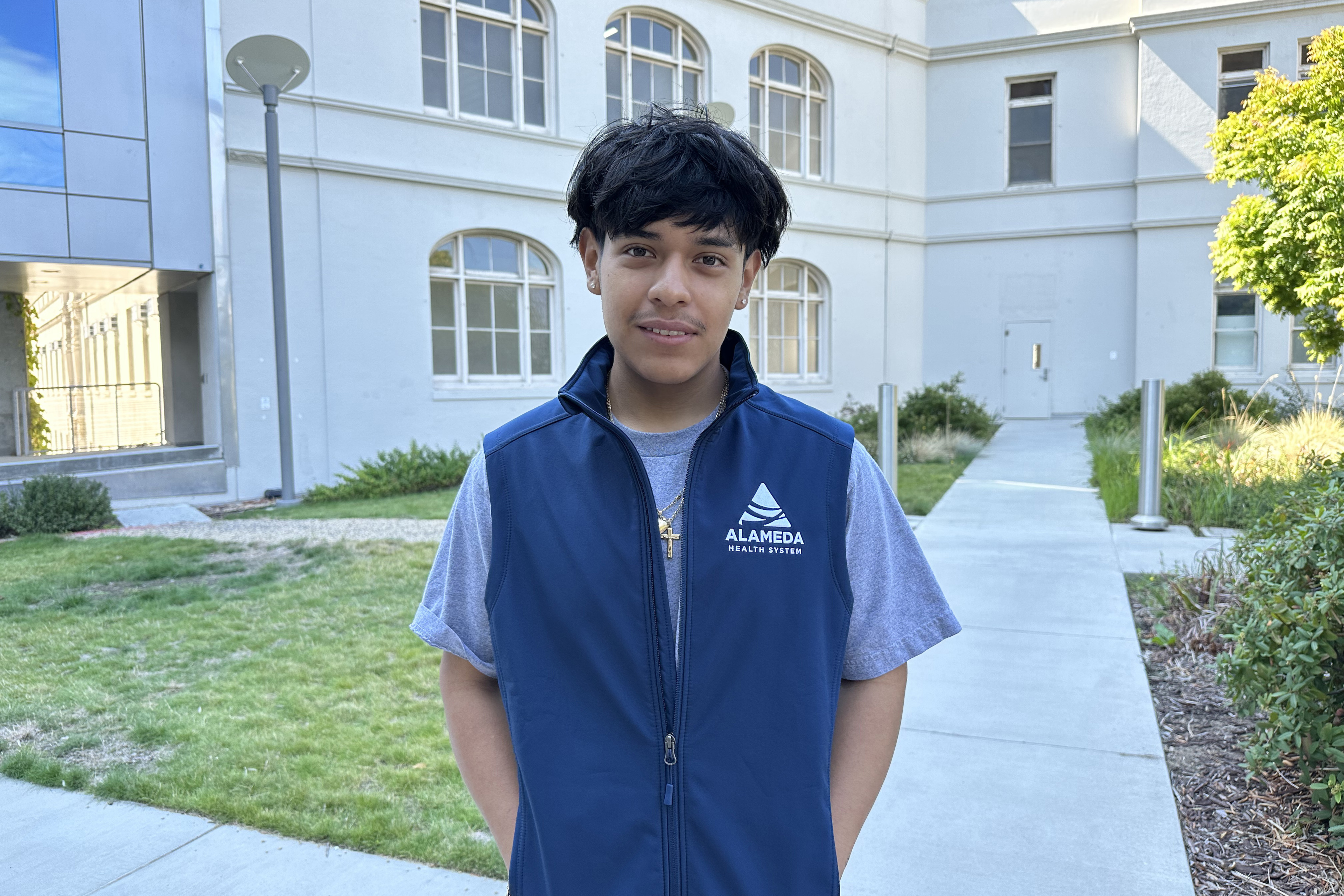 A young man wearing a vest with the words "Alameda Health System" on it stands outside of a building and looks at the camera.