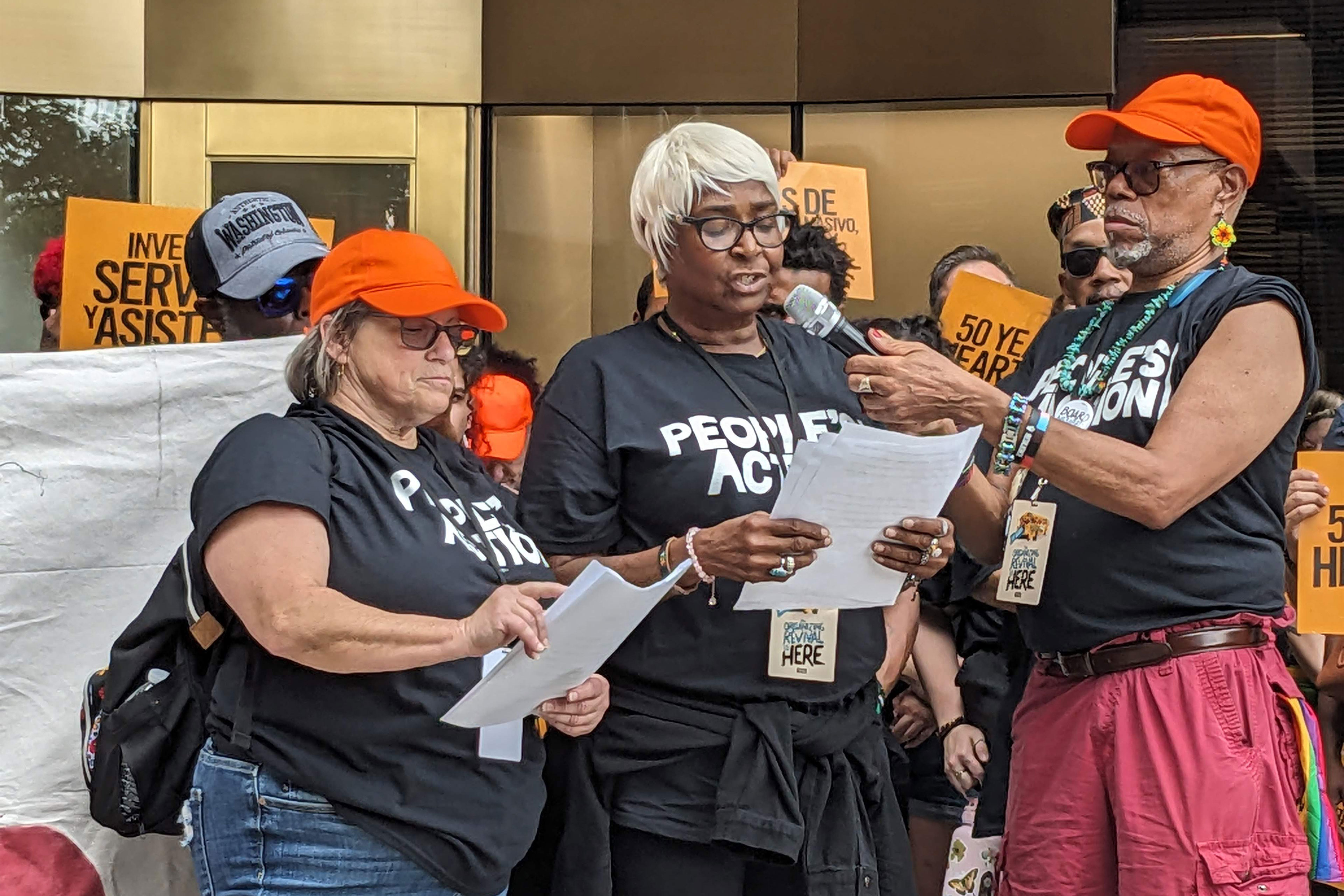 A woman stands between two people and reads from piece of paper. The person to her left holds the microphone for her to speak into.
