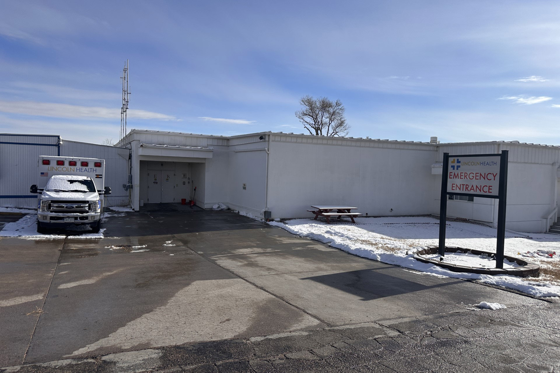 The photo shows the exterior of Lincoln Health. A ambulance covered in snow is parked on the left of a paved driveway. A sign that reads, "Lincoln Health / Emergency Entrance" stands to the right in a grassy, snow-covered area.