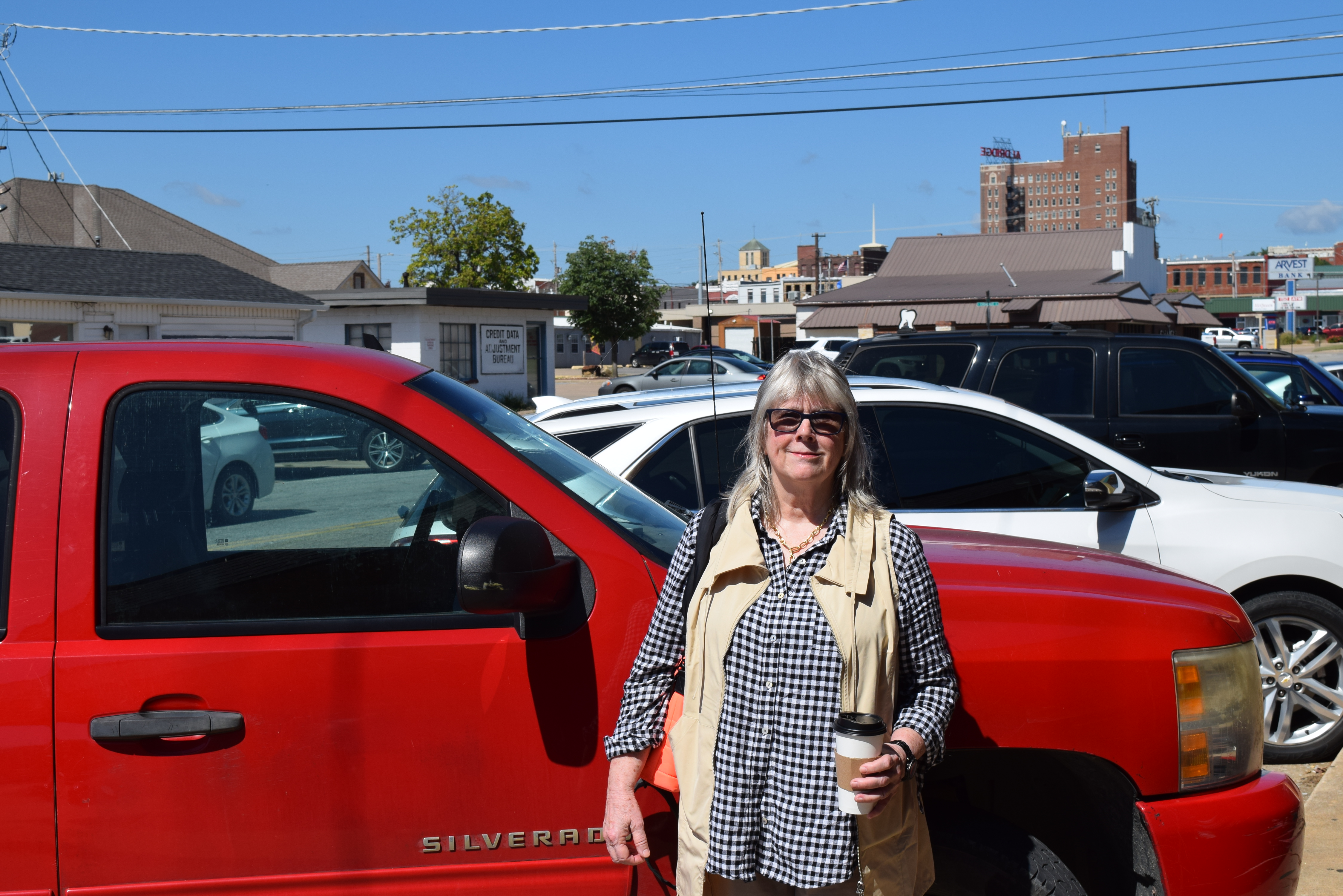 A woman in a black and white top holds a single use coffee cup and stands in front of a red SUV truck.