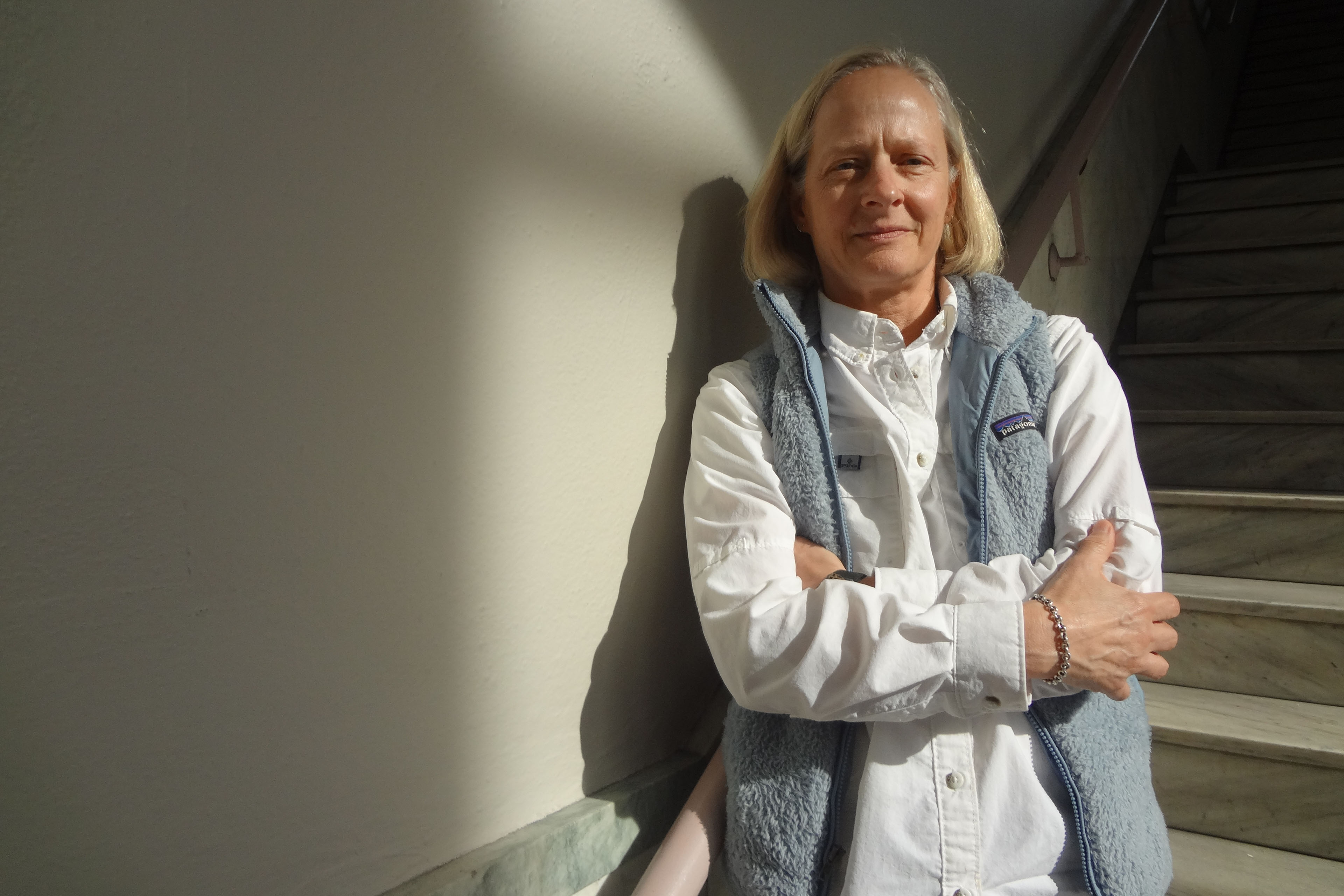 Jan Rader stands beside a stairwell with her arms crossed as she looks towards the camera.