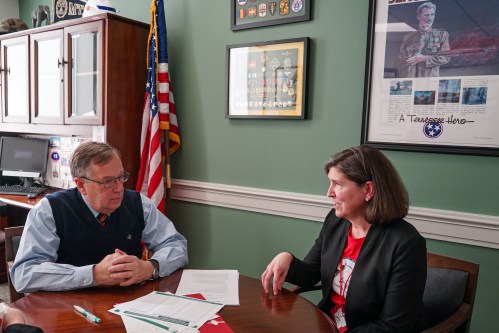 Barb Dentz (right), sits beside state representative, Sam Whitson (left), in an office room. The walls are an olive green and are adorned by framed medals. An American flag stands in the corner, behind them.