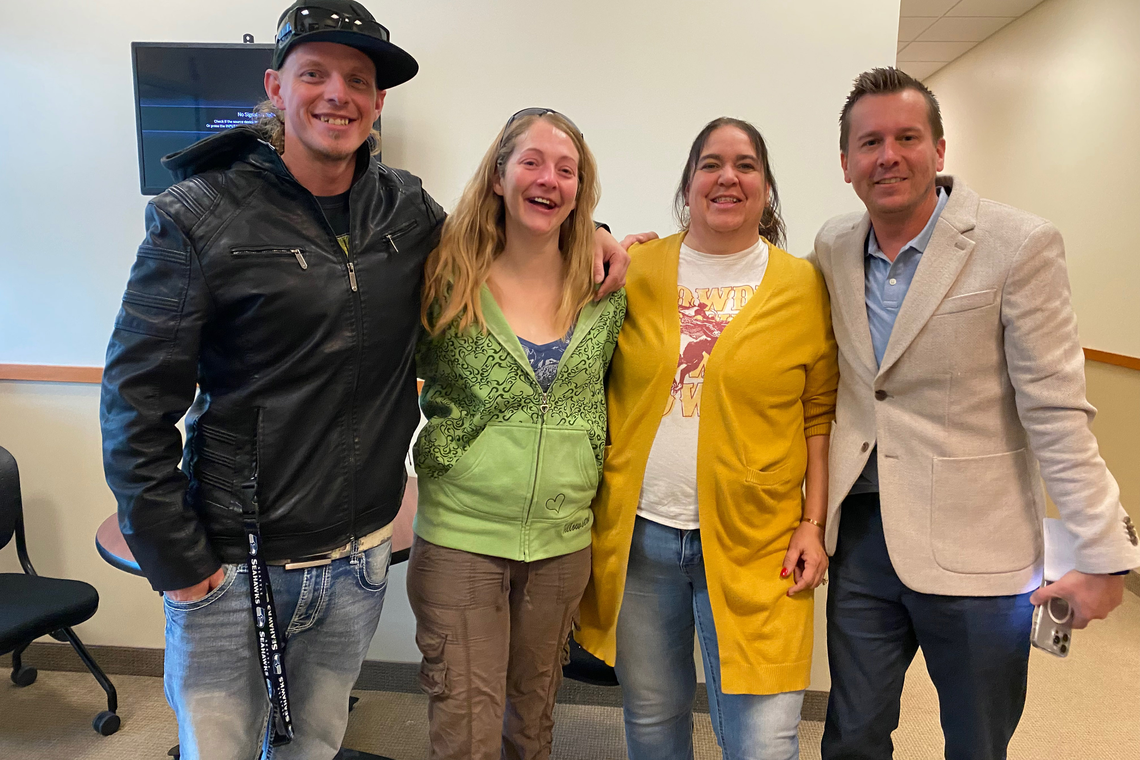 From left to right: Garrett Clark, Shantel Clark, Cheryl Swapp, and Ryan Montag. They stand in a row, smiling towards the camera, in a room with blank walls and fluorescent overhead lighting.