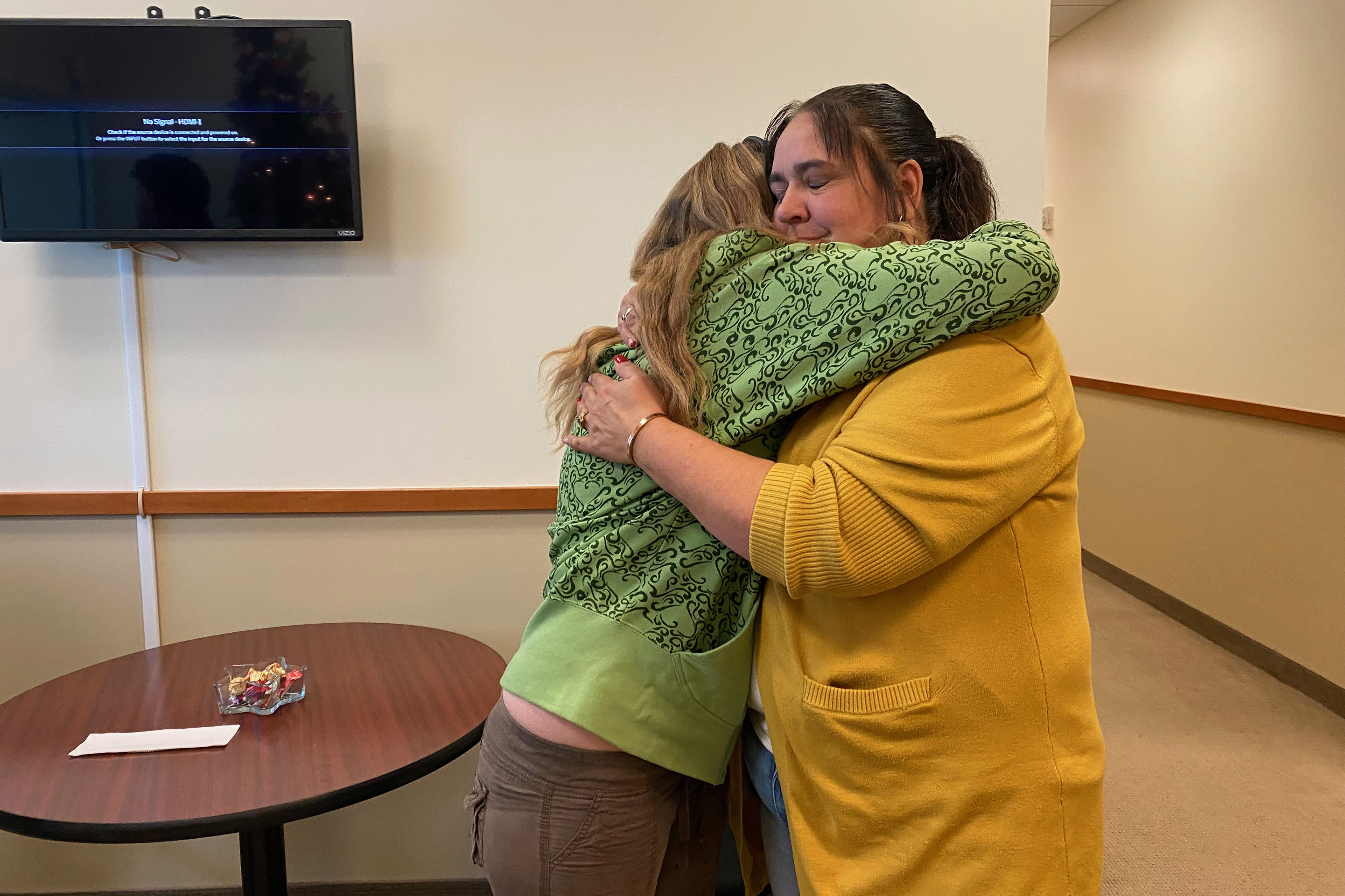 Shantel Clark hugs Cheryl Swapp in a room with blank walls and fluorescent overhead lighting. Clark wears a green sweatshirt with a black ornate pattern. Swapp wears a yellow sweater.