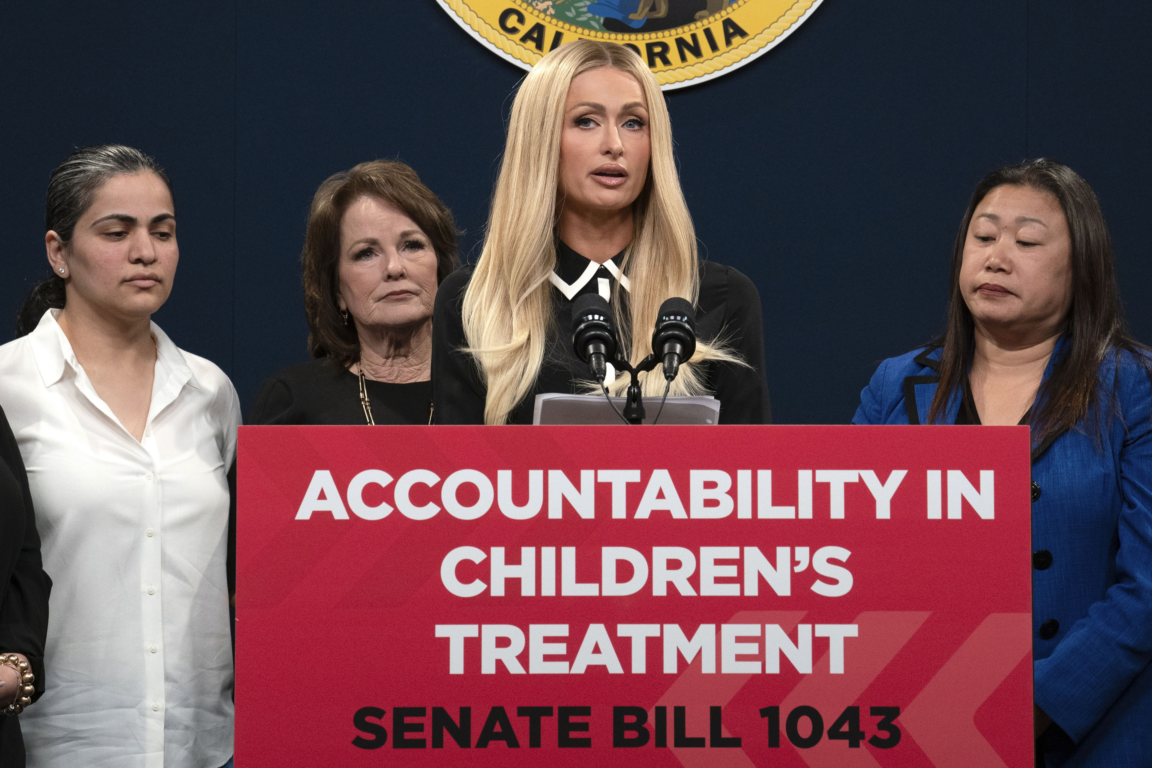 A tall, blonde woman in her early 40's stands a podium. The podium has a red sign that reads, "ACCOUNTABILITY IN CHILDREN'S TREATMENT / SENATE BILL 1043"