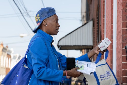 A woman in a backwards blue cap and matching blue sweat jacket hands out Narcan door to door