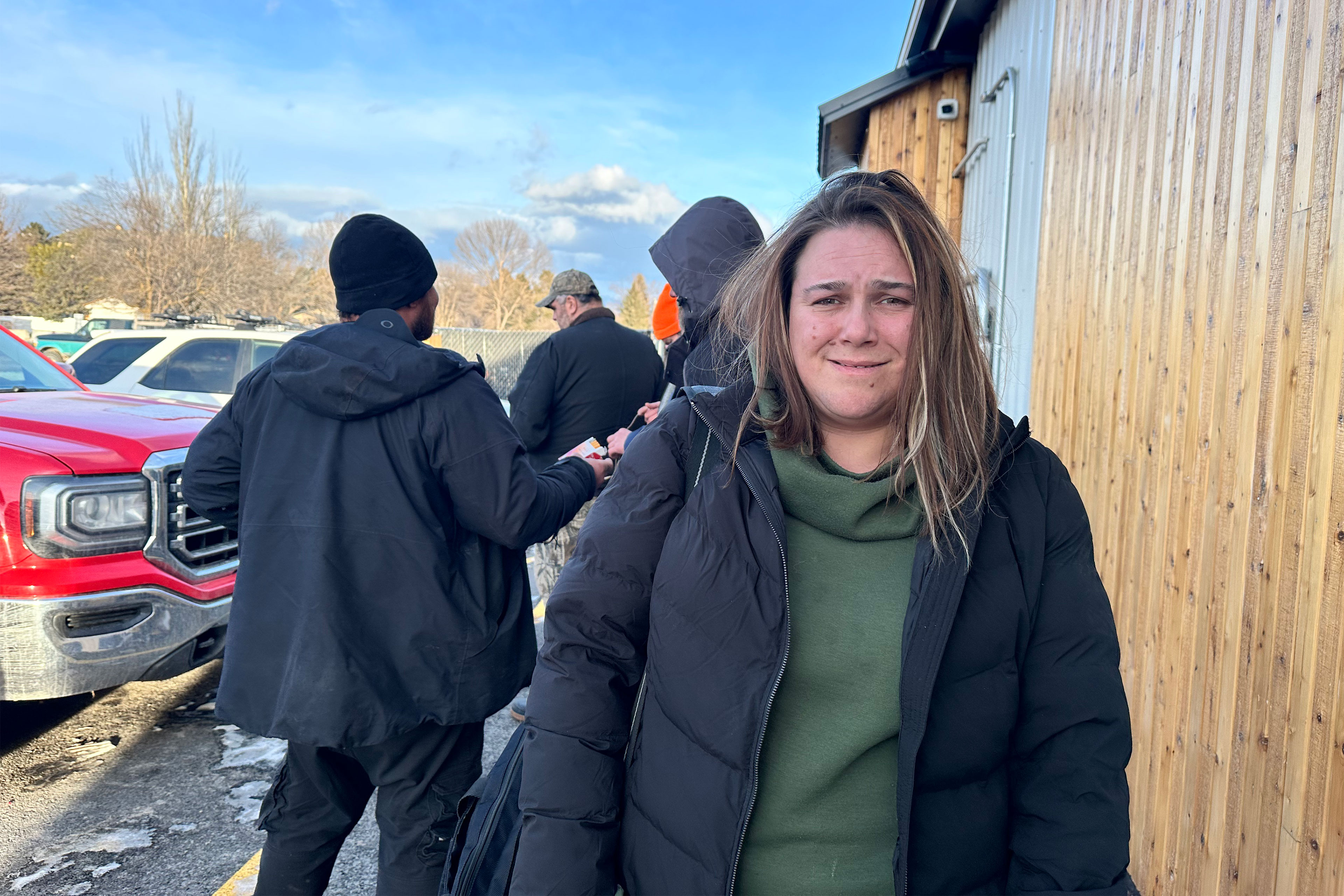 A woman wearing a green sweatshirt under and black puffy jacket stands and smiles at the camera.