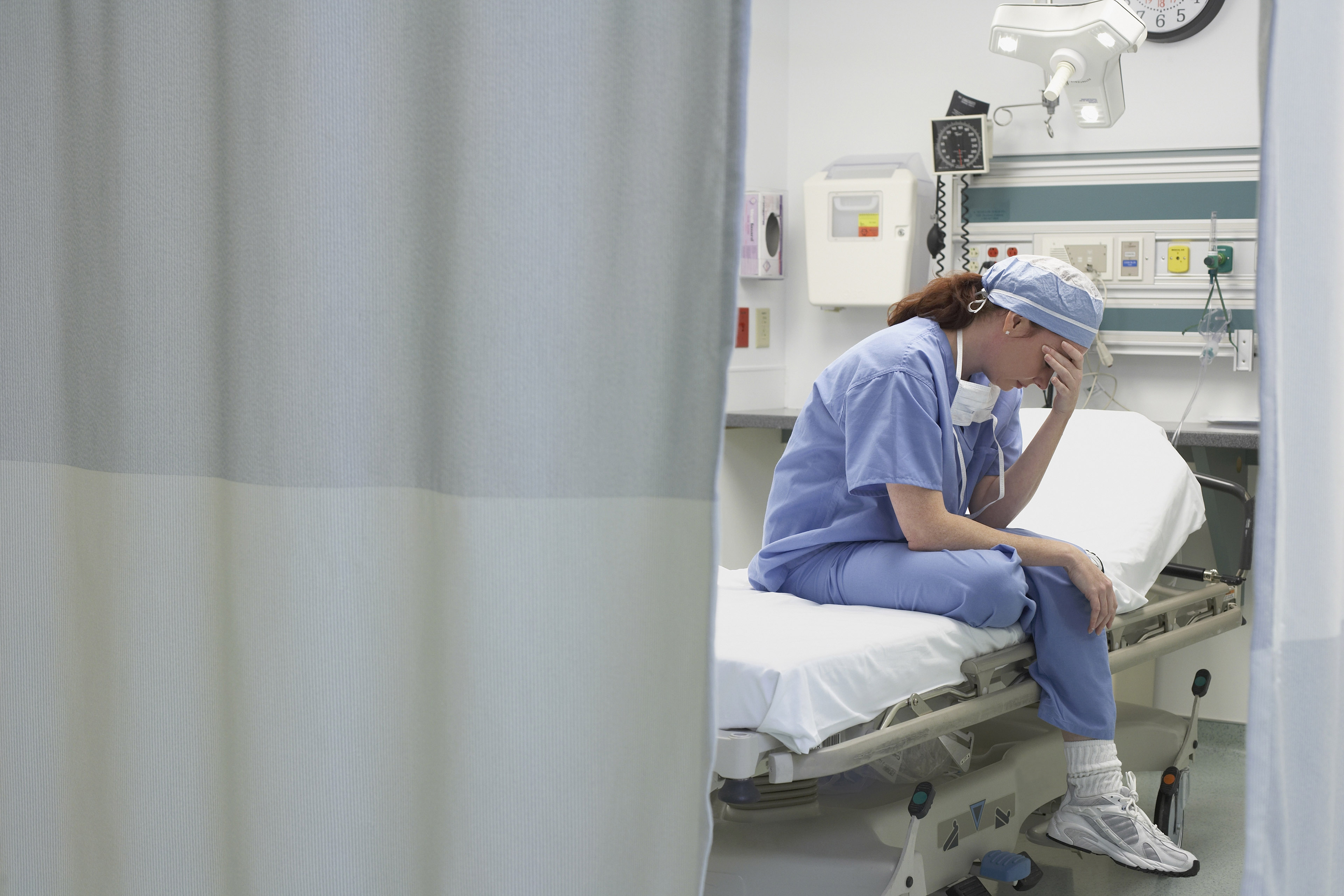 A female doctor sits with her head in hand hand on an empty hospital bed.