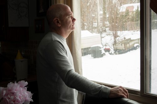A side-view of a man standing at a window and looking out. His right had rests on the window sill.