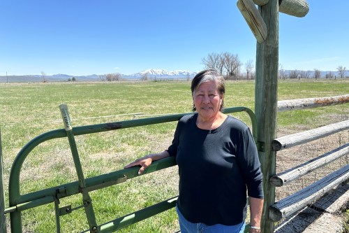 Myrna Broncho is standing outdoors beside a wooden fence that lines a large, open field on a sunny day.