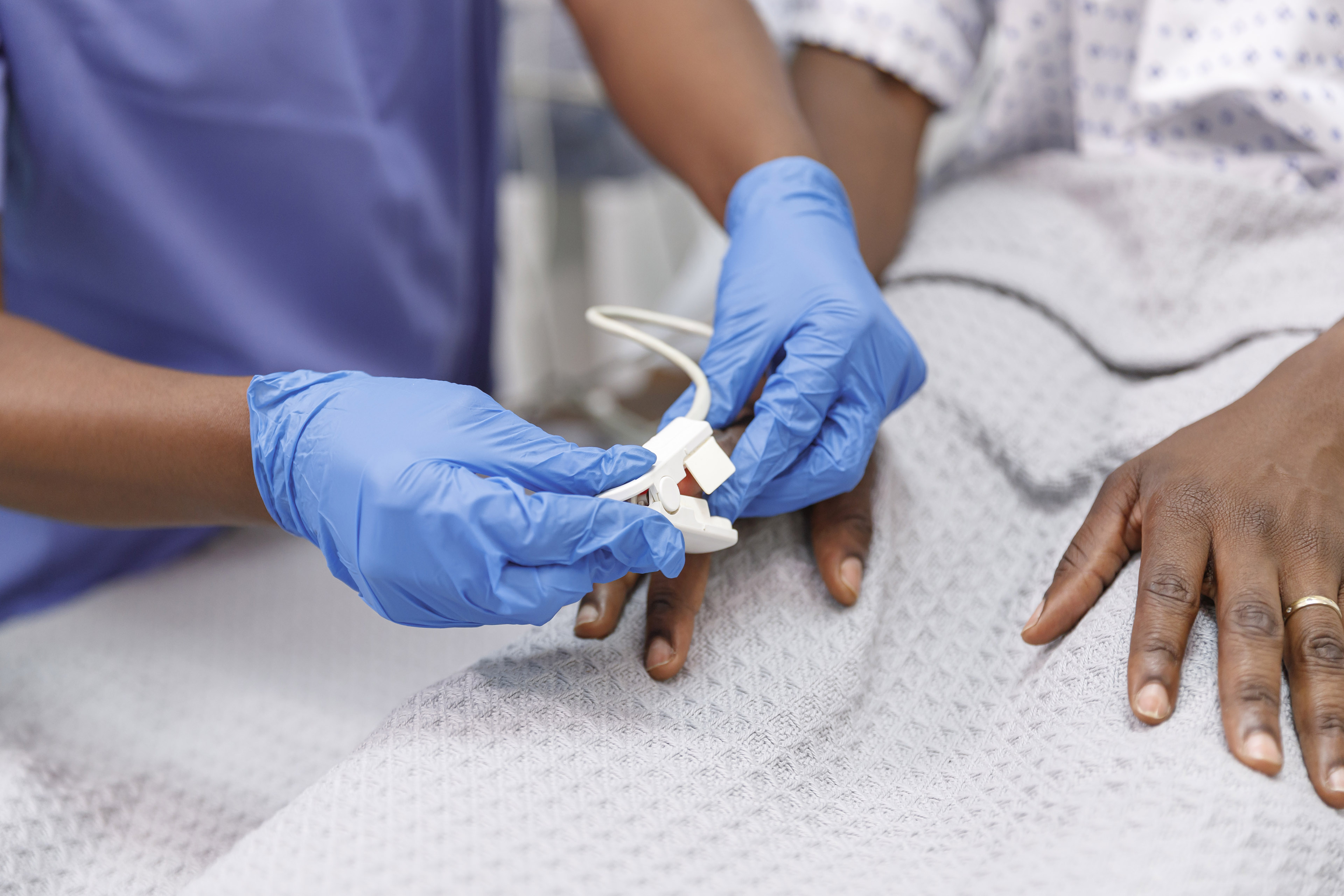 A close-up photo of a medical professional placing a pulse oximeter on the finger of a hospitalized patient who is lying in bed.