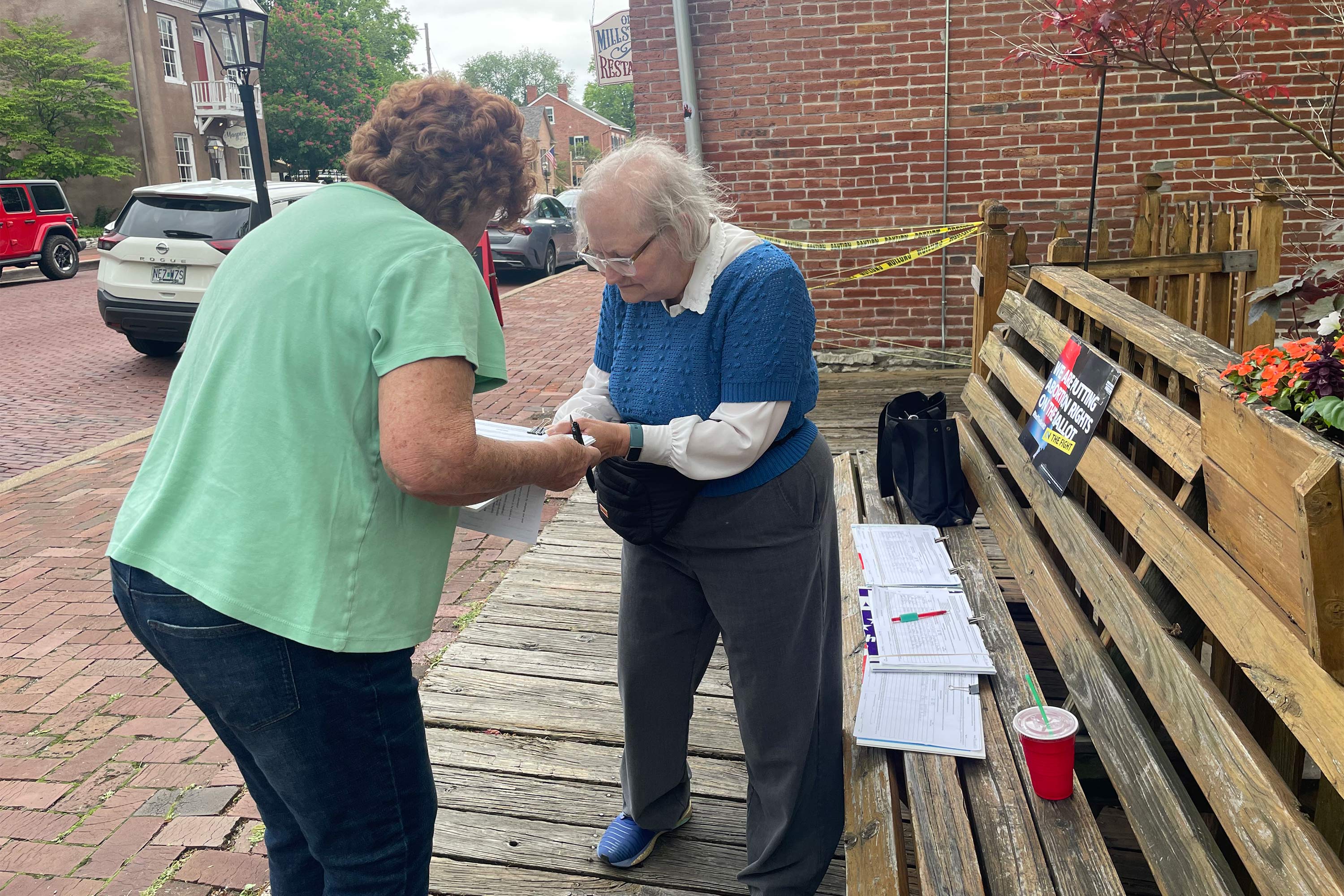 A photo of two women standing outside. The one on the left holds a stack of papers and a pen.