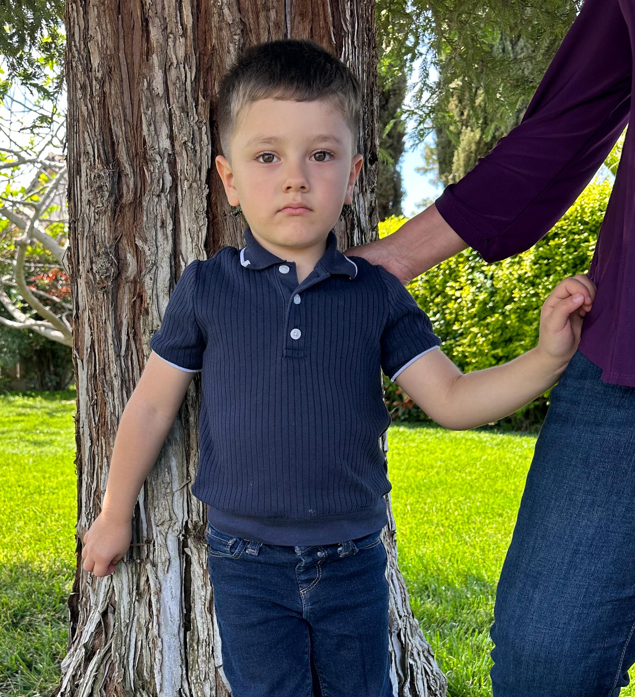 A photo of a young boy standing outside by a tree, holding on to his mother's shirt.