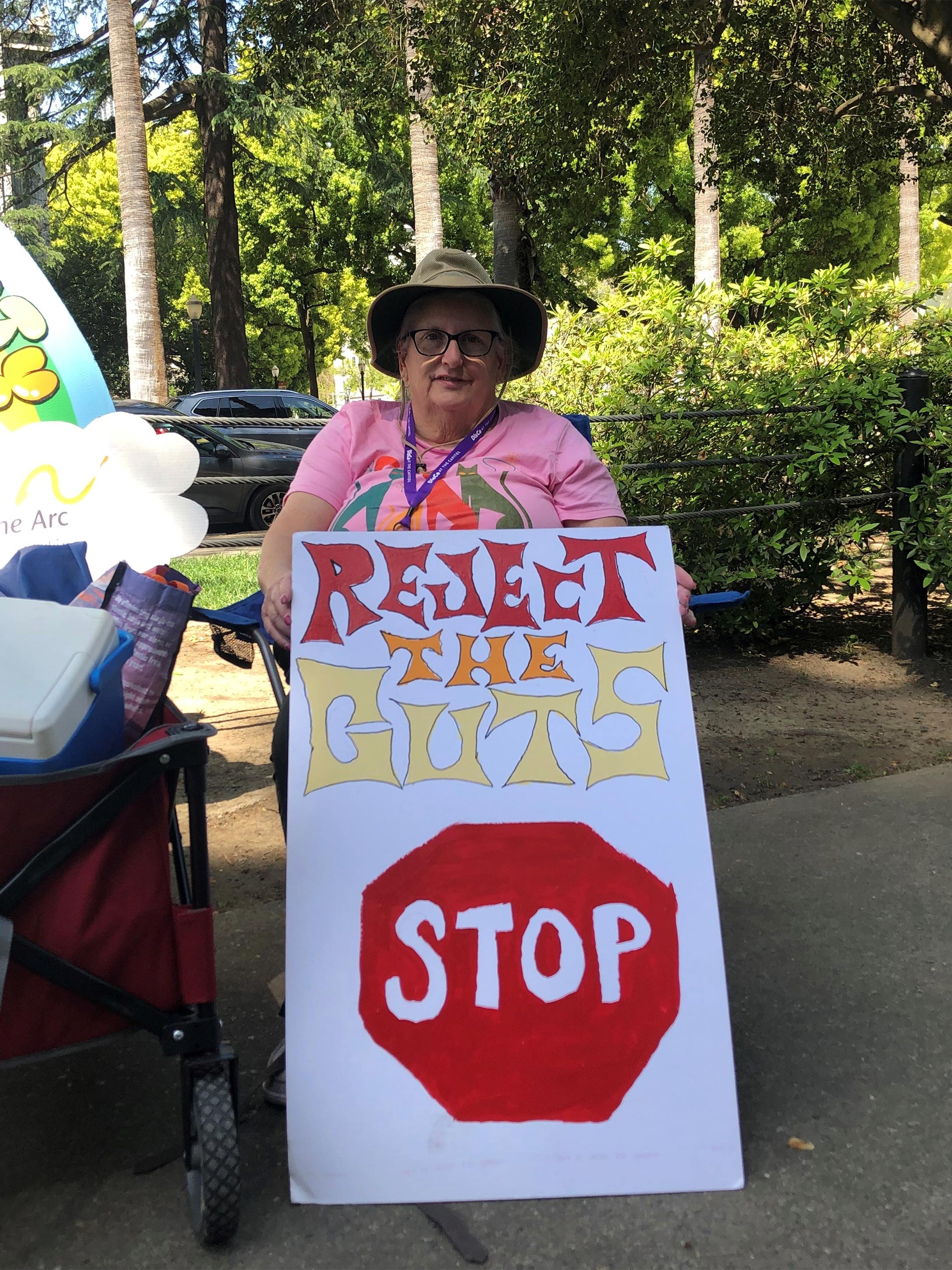 A photo of an activist sitting outside with a sign that reads, "Reject the cuts."