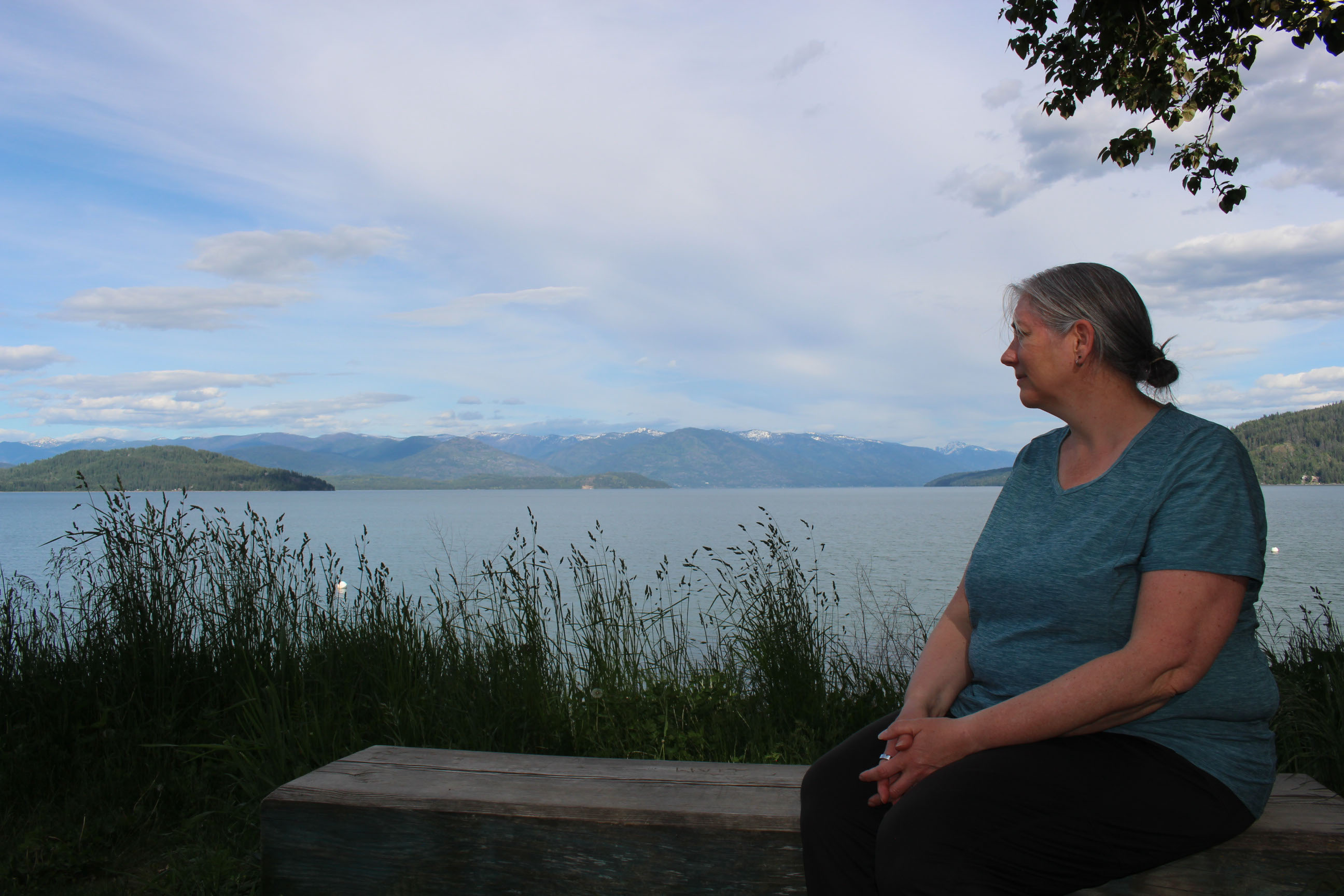 A portrait of Jonell Anderson, who is sitting on a wooden bench by a large body of water on a partly-sunny day.