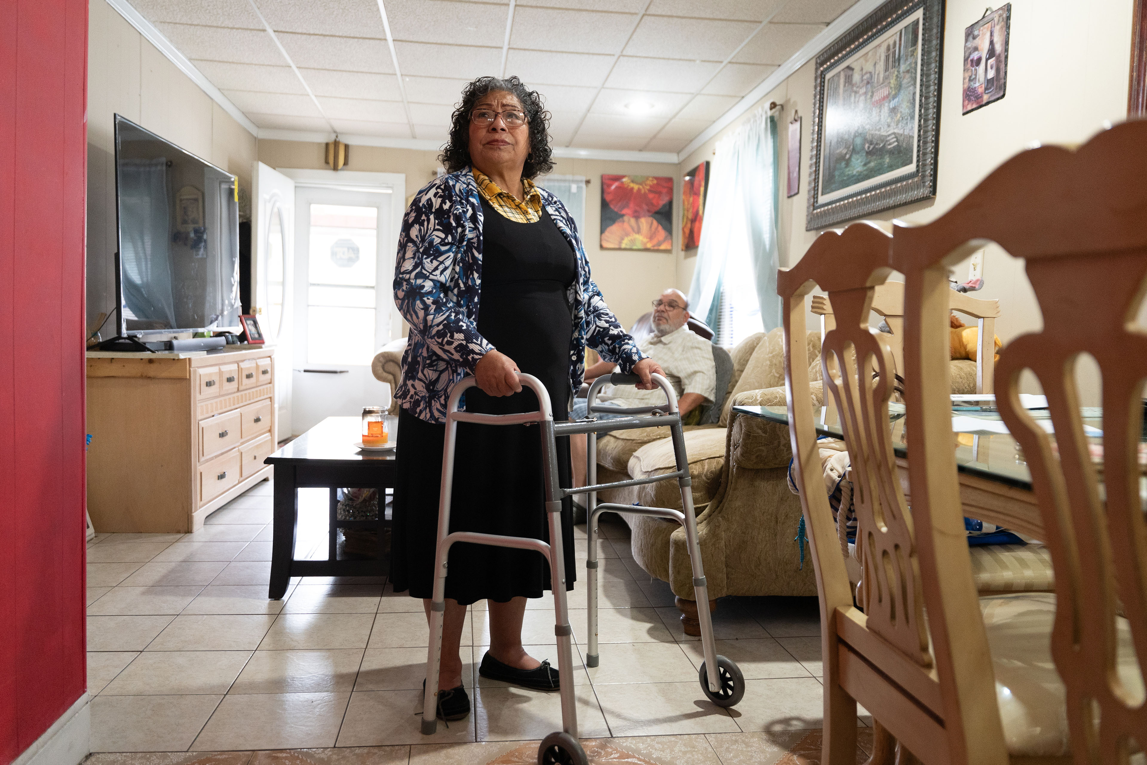 A woman wearing a black dress and blue and white shirt, holding on to a walker, stands next to a dining room table with a man sitting on a couch in the background