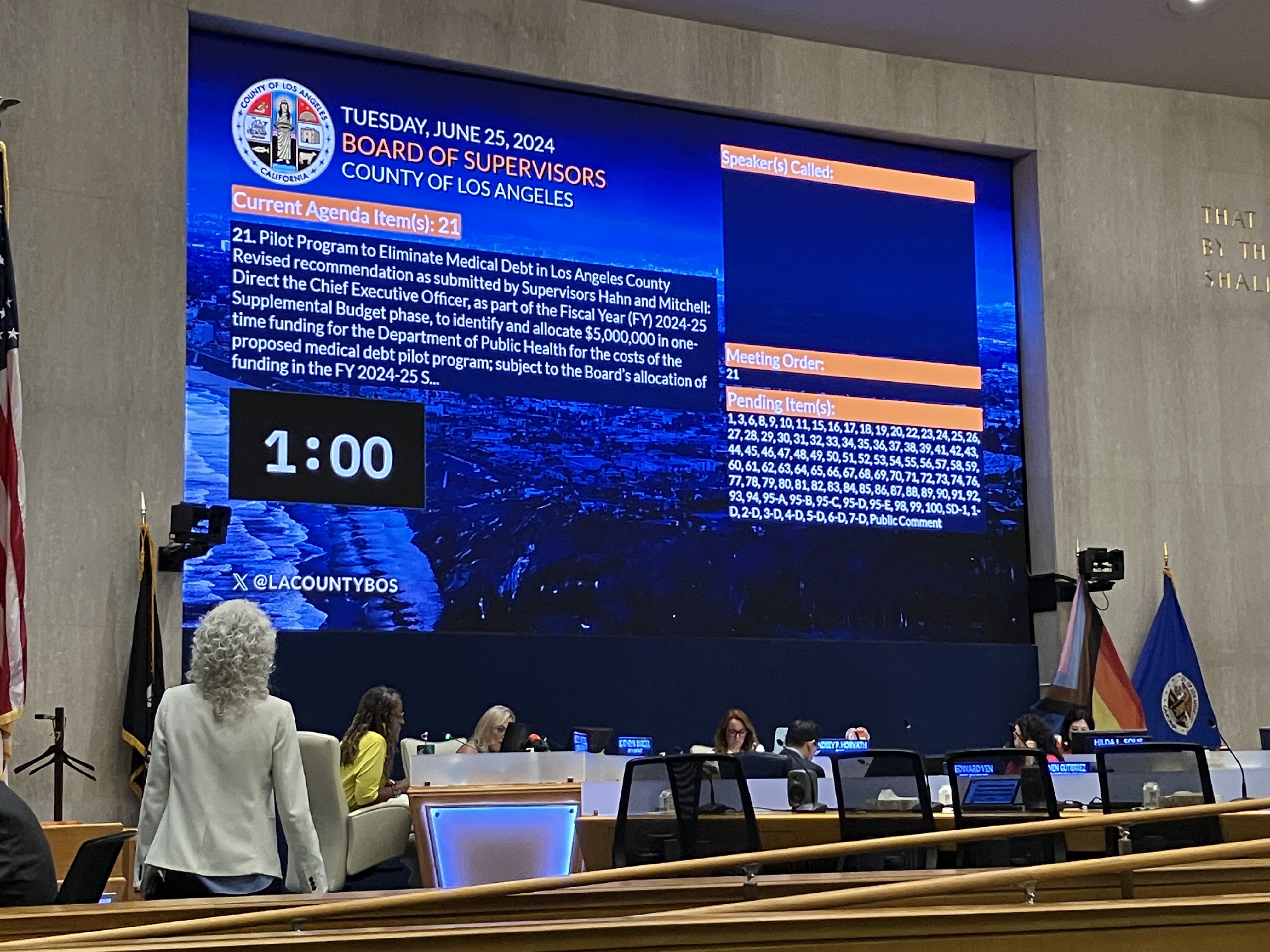 A projector screen says, "Tuesday, June 25, 2024 / Board of Supervisors / County of Los Angeles / Current Agenda Item(s): 21 / 21. Pilot Program to Eliminate Medical Debt in Los Angeles County Revised recommendation as submitted by Supervisors Hahn and Mitchell: Direct the Chief Executive Officer, as part of the Fiscal Year (FY) 2024-25 Supplemental Budget phase, to identify and allocate $5,000,000 in one-time funding for the Department of Public Health for the costs of the proposed medical debt pilot program; subject to the Board's allocation of funding in the FY 2024-25 S..."
