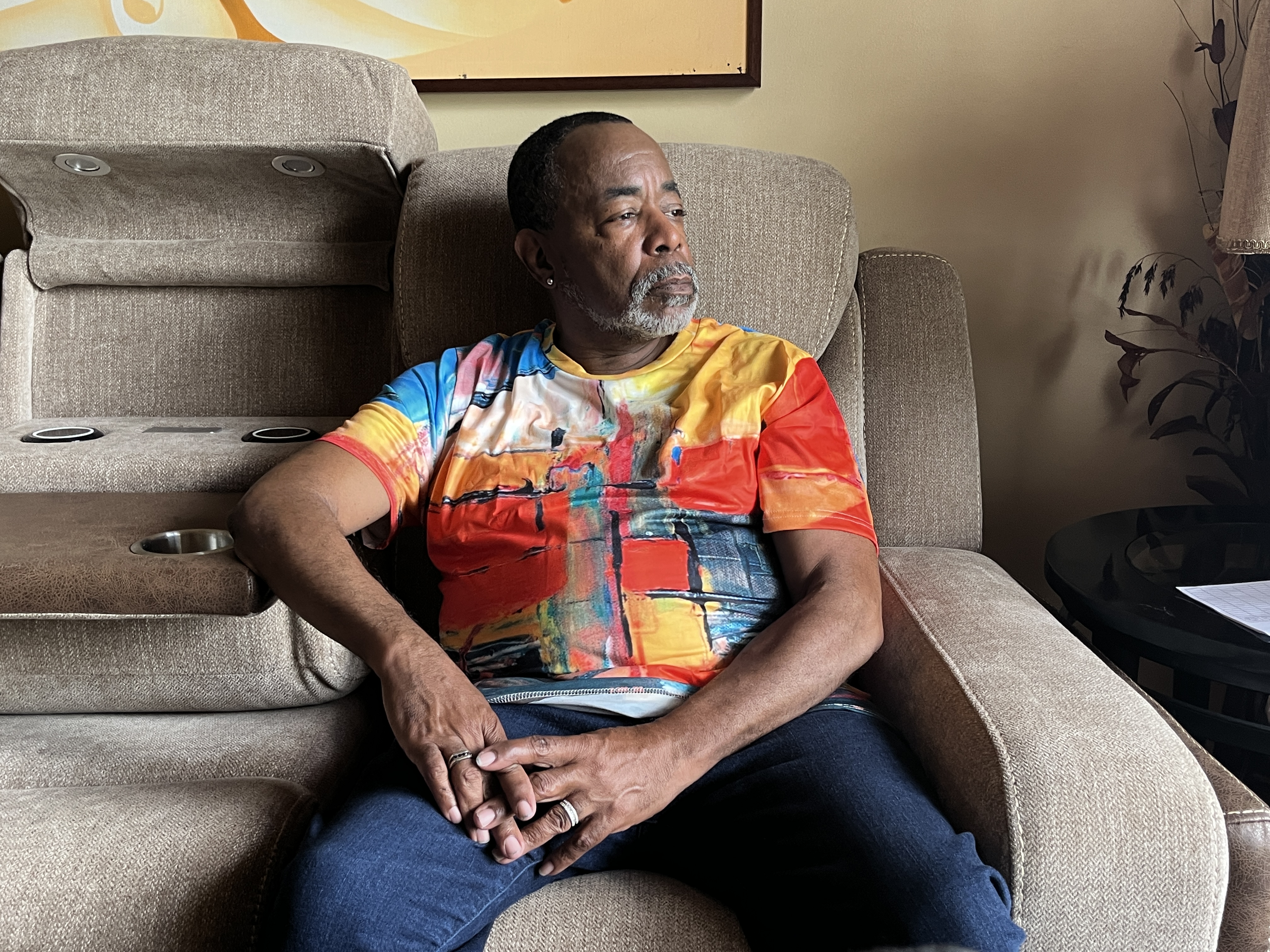 A man in a color t-shirt sits on a brown couch, looking out the window to his left