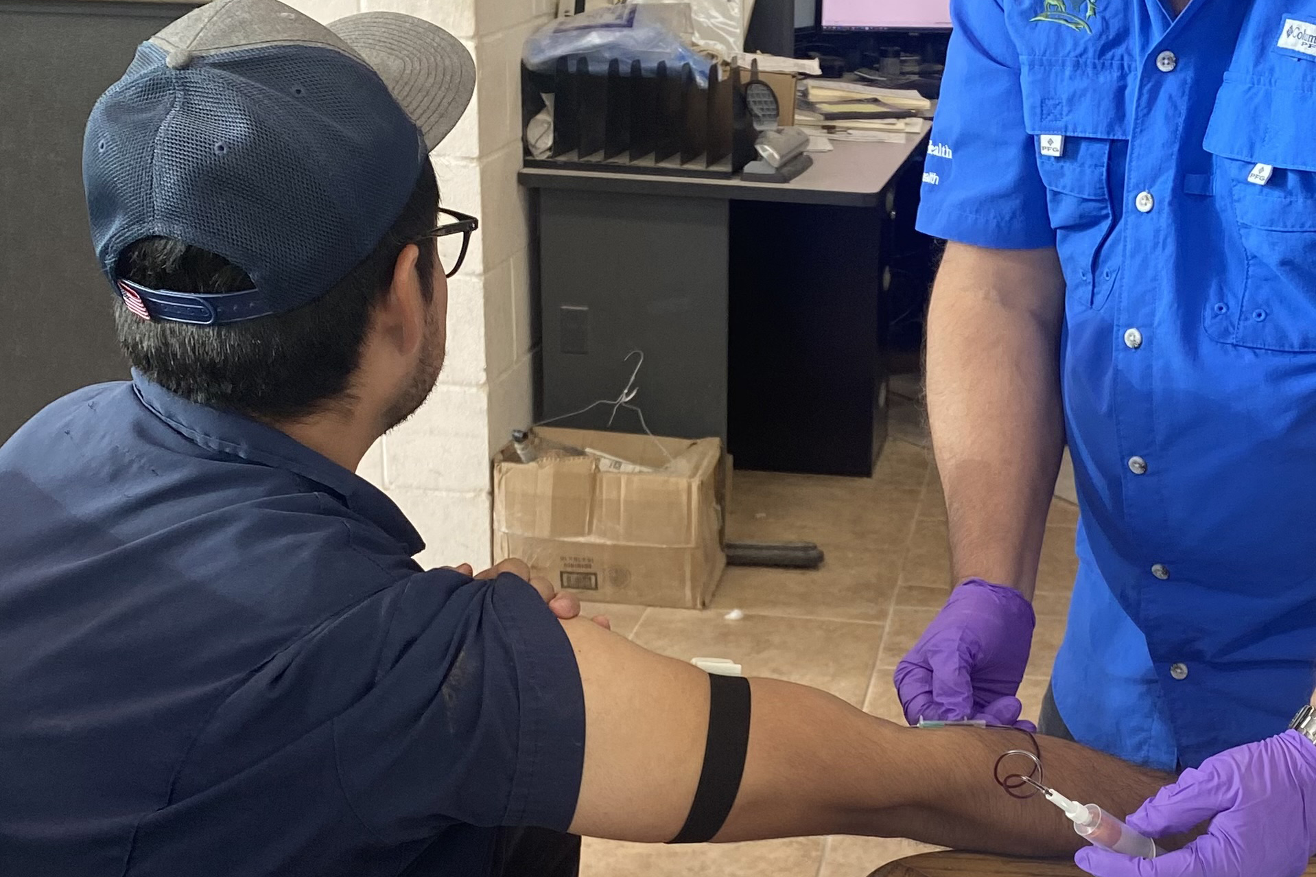 A man in a dark blue shirt and cap faces away from the camera and stretches out his right arm where another person wearing purple medical gloves draws blood.