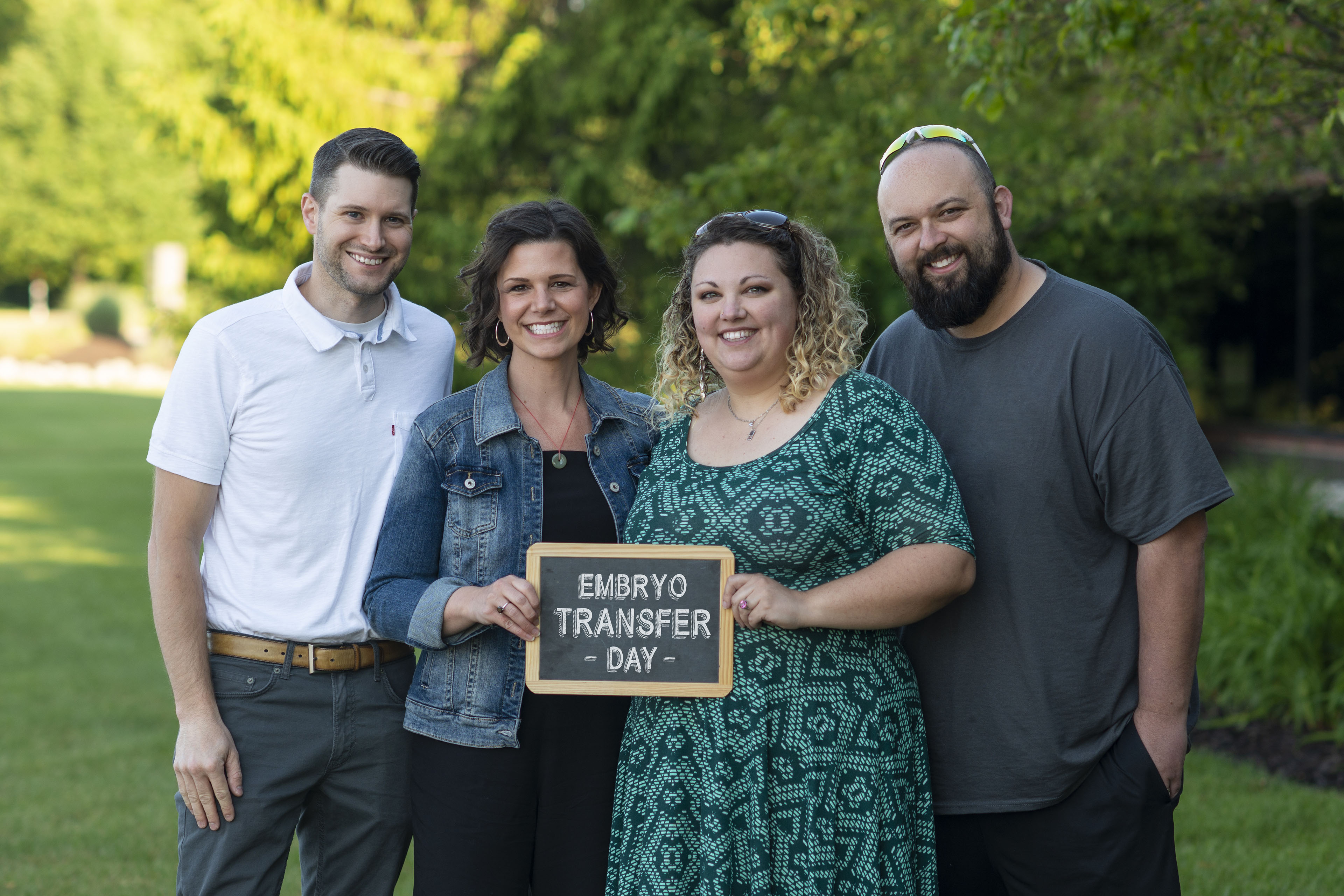 Four people, two men and two women, pose for a portrait holding a sign that says "embryo transfer day"