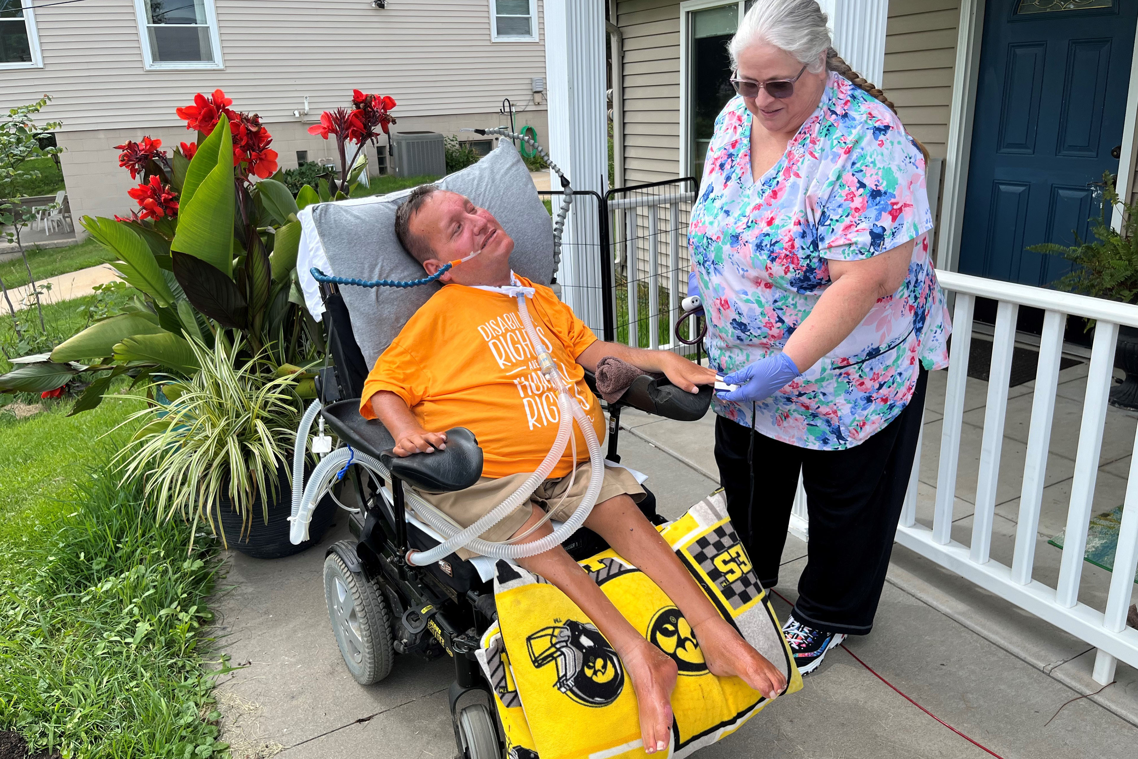 A nurse stands beside a man in an electronic wheelchair outside of a suburban home.