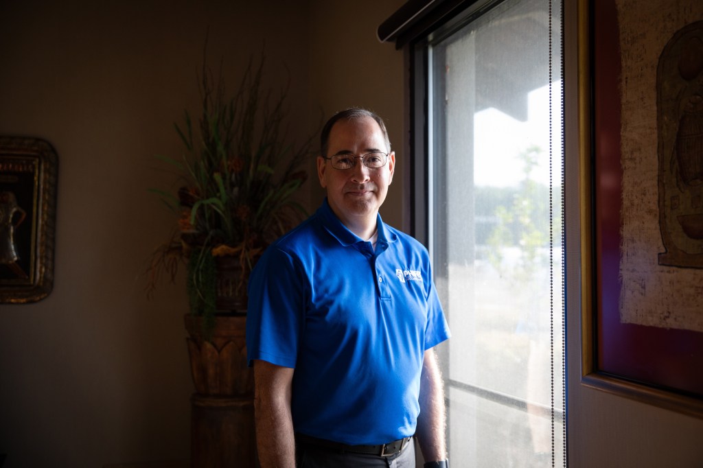 A man in a blue polo shirt stands in front of a window