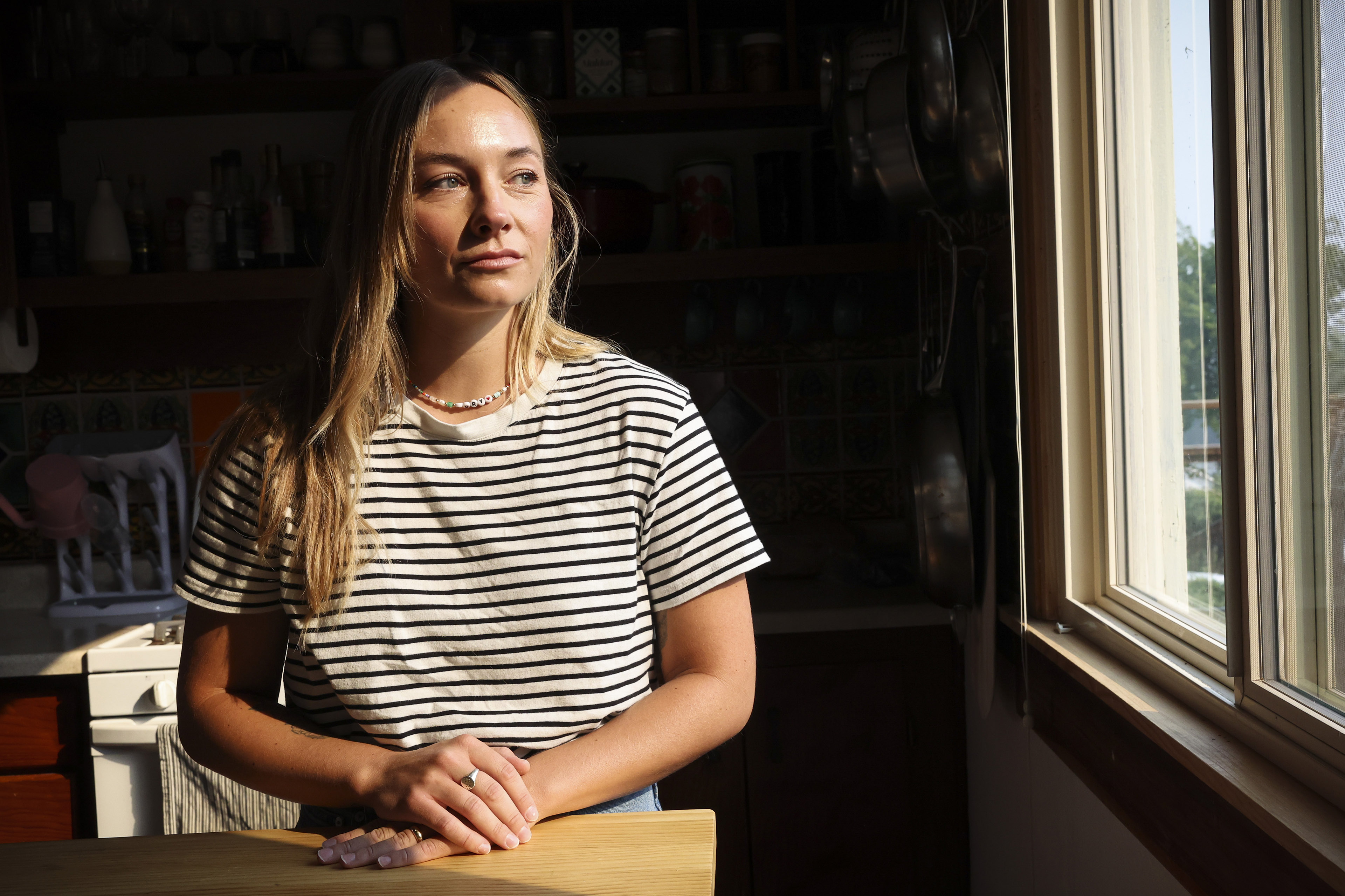 A woman with blond hair and a black and white striped shirt stands in the sun in front of a kitchen window