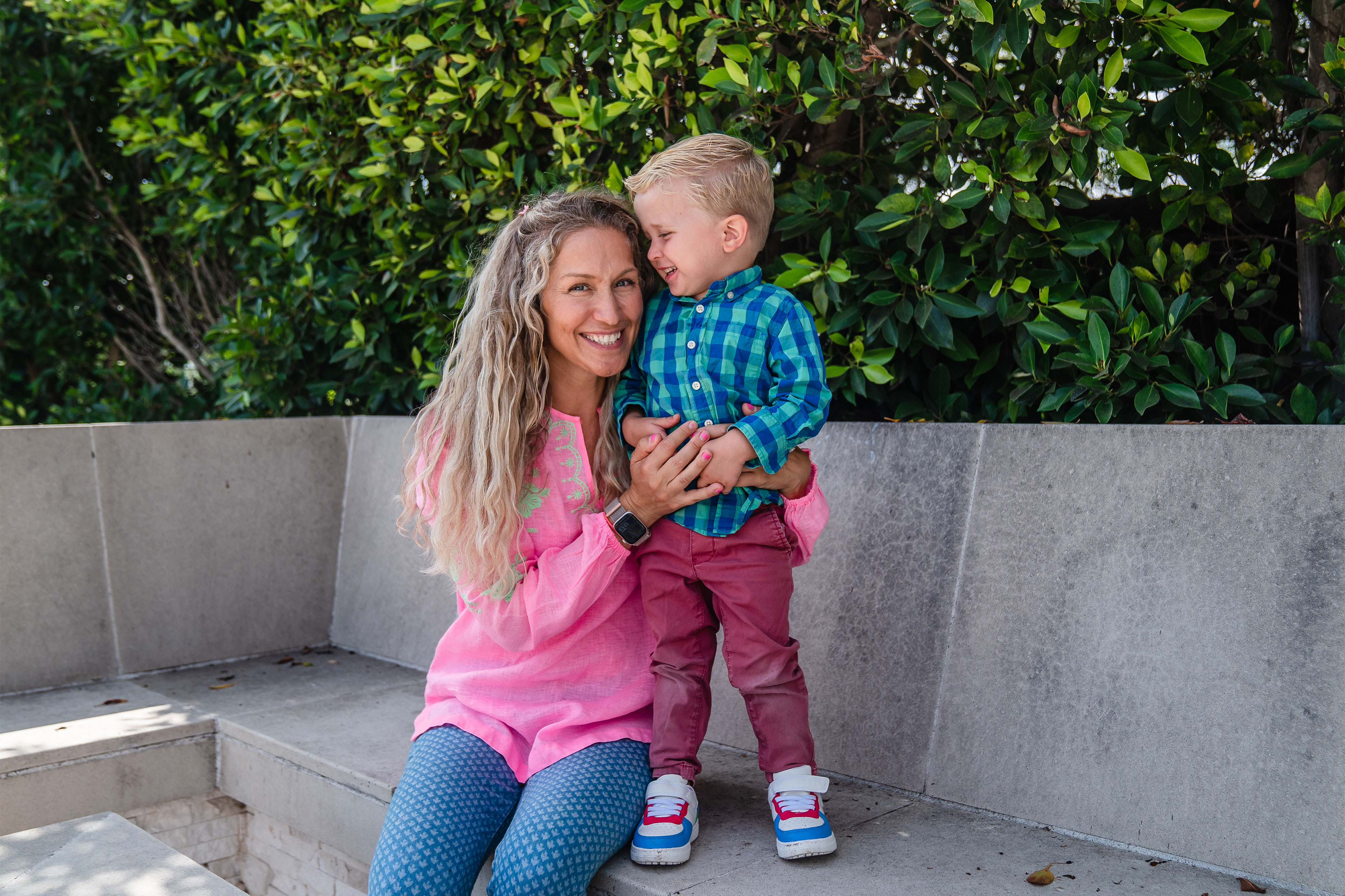 A woman sits on a concrete bench with her arm around a young boy standing on the bench beside her. She smiles at the camera and he smiles at her.