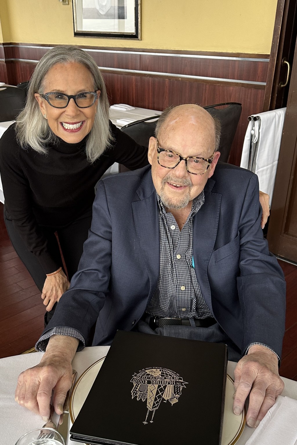 Joy Loverde, an older woman with shoulder length gray hair and glasses, stands beside Gardner Stern, a senior man wearing glasses and a suit jacket. They are sitting down at a table with a white tablecloth.