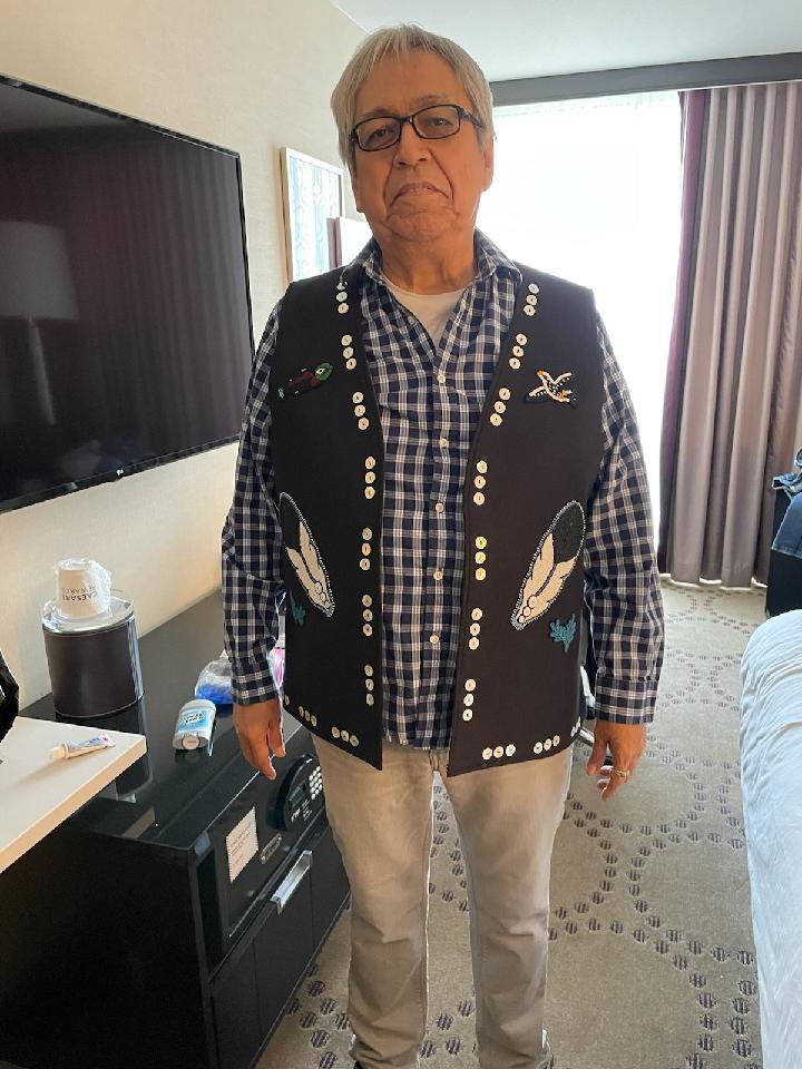 A man wearing a vest with animal and feather designs on it over his checkered shirt stands and looks at the camera.