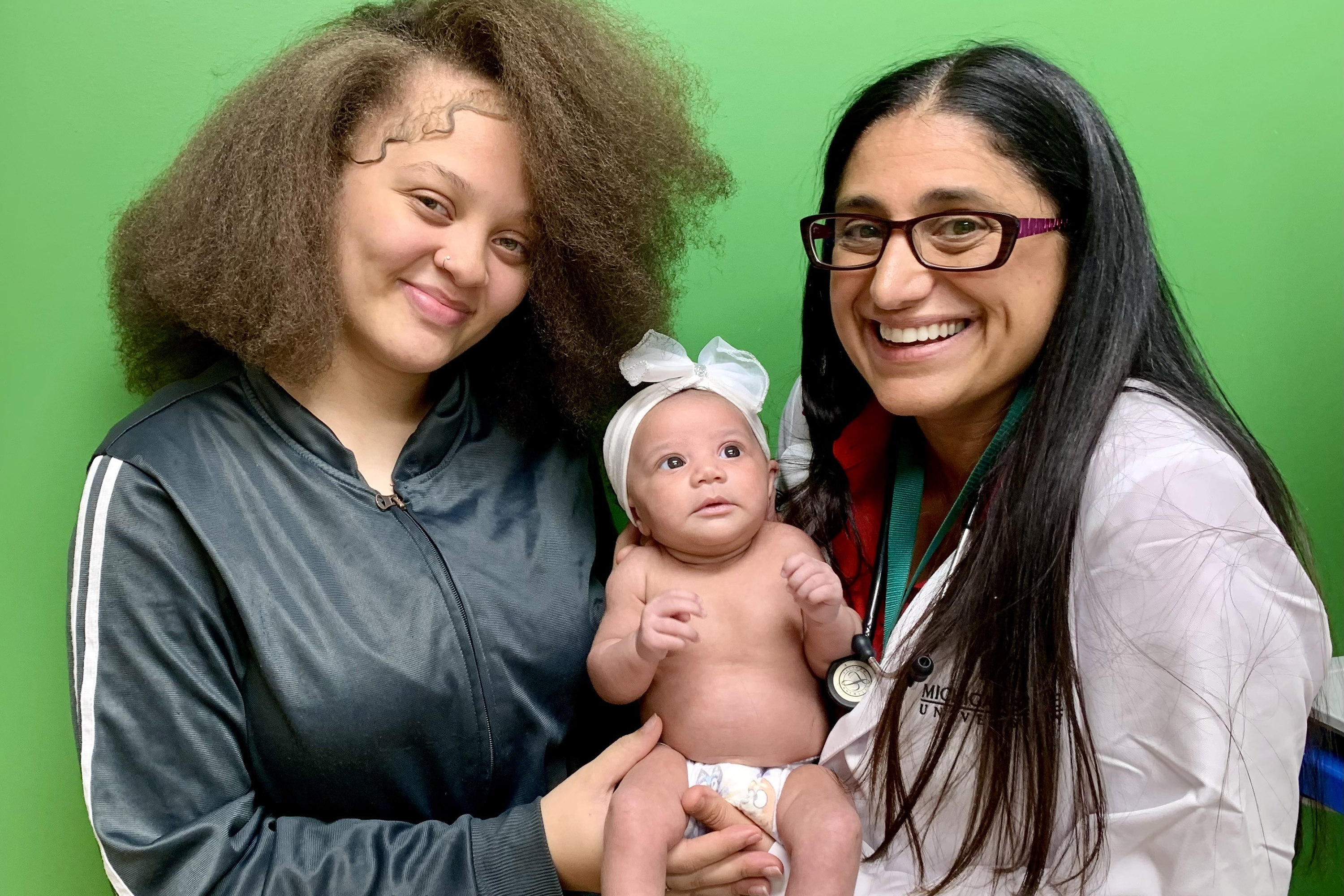Two women smile at the camera while holding a baby with a white bow on her head