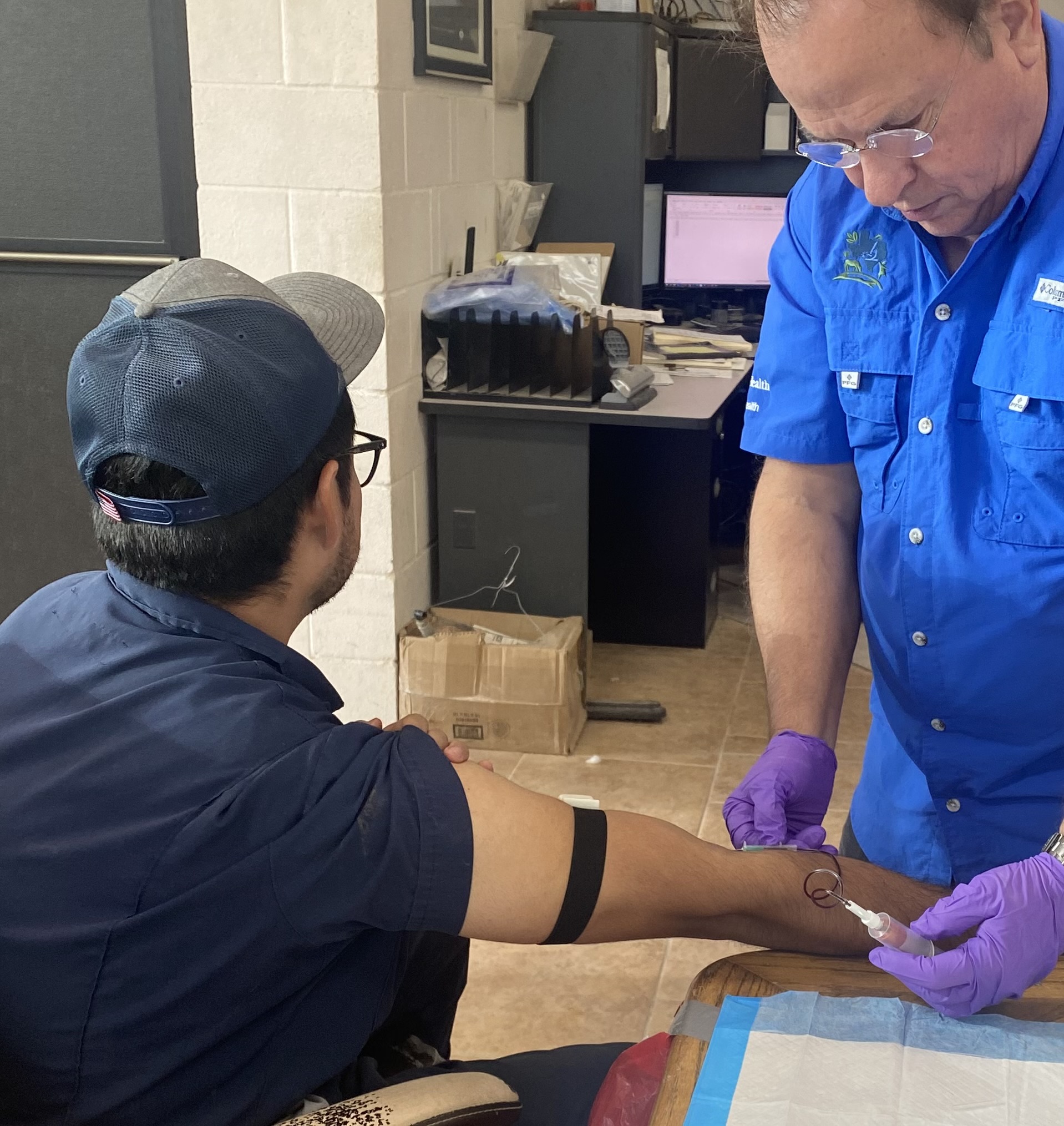 A man in a dark blue shirt and cap faces away from the camera and stretches out his right arm where another person wearing purple medical gloves draws blood.