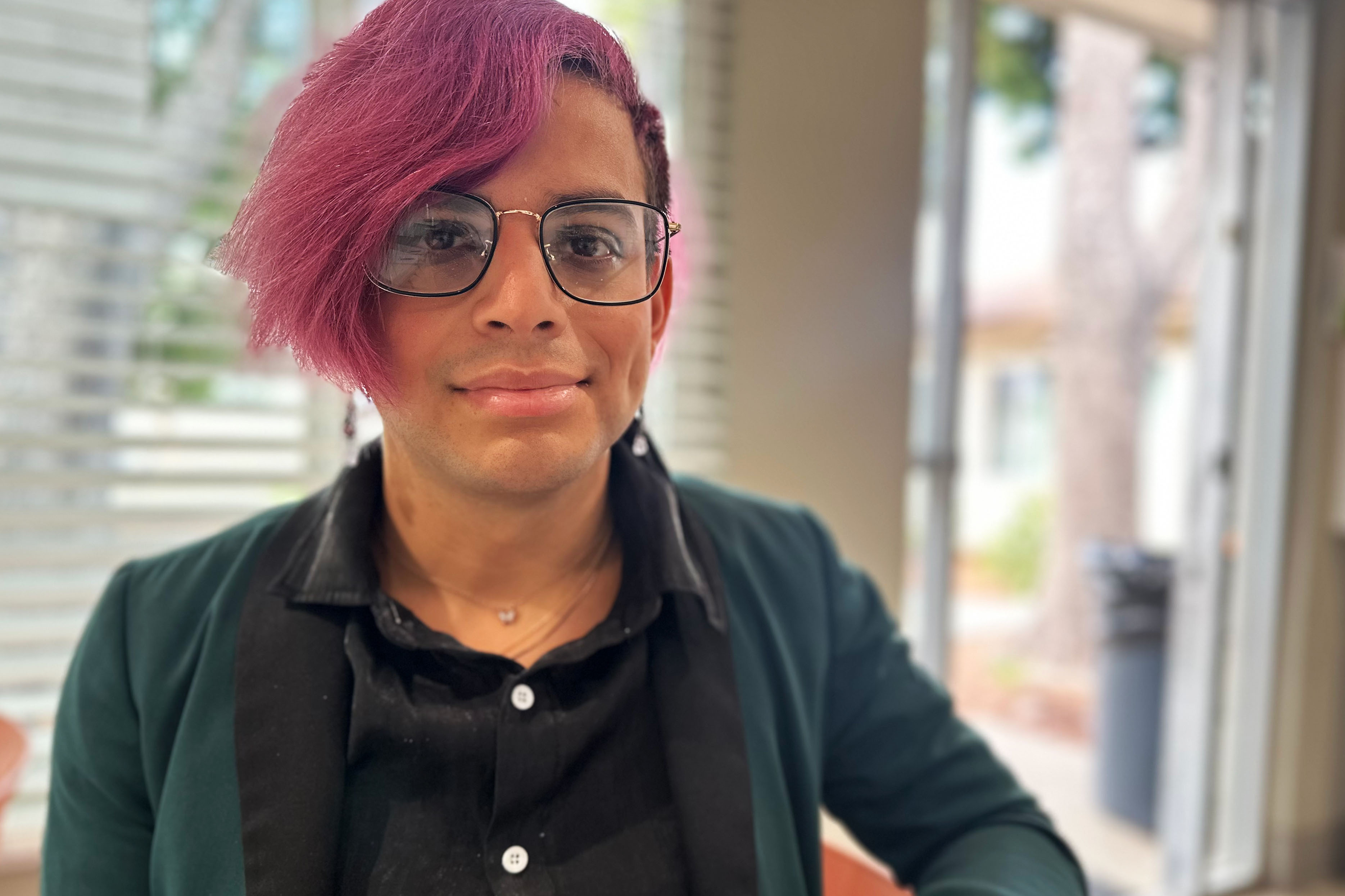 An adult woman with short, pink hair and glasses looks towards the camera with a subtle smile.