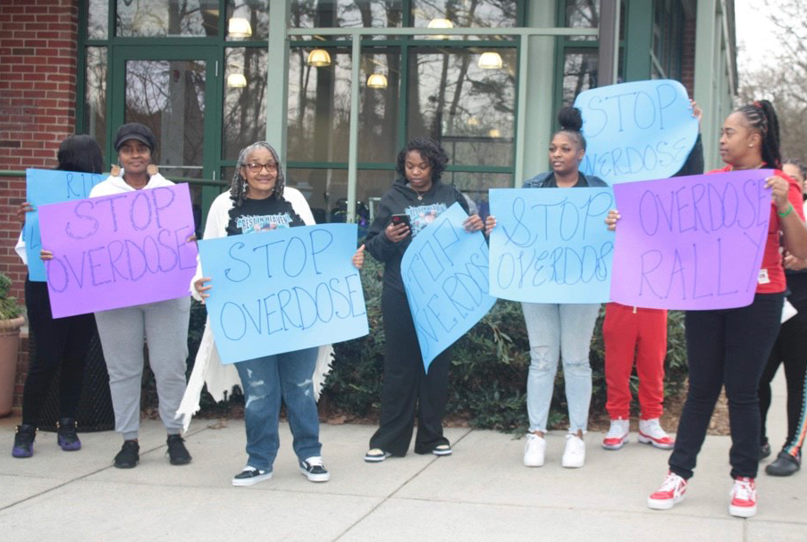 A photo of a group of people holding signs in front of a building. Most of the signs read, "Stop overdose," and one reads, "Overdose rally."