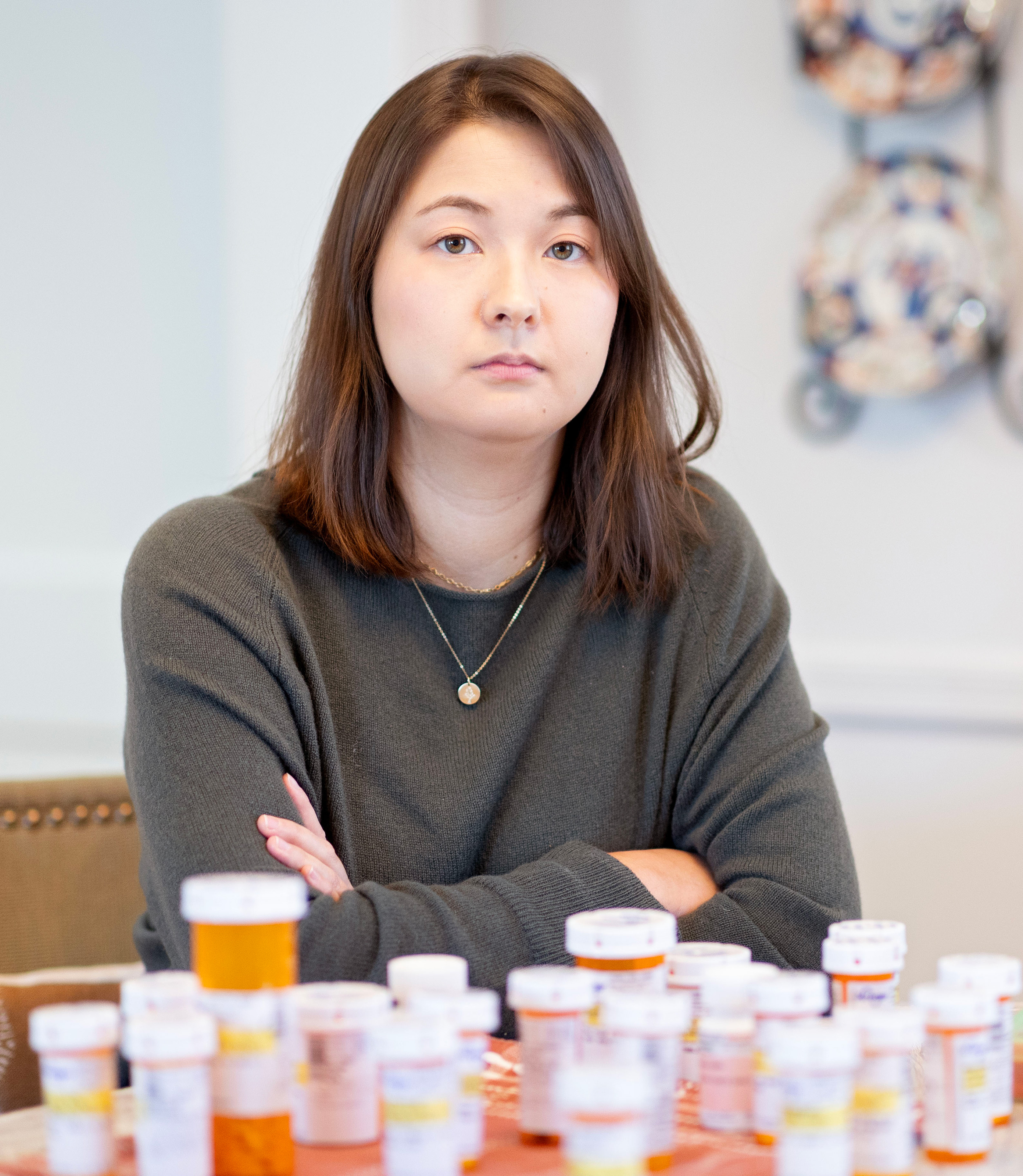 A photo of a woman sitting at a table with over a dozen pill bottles in front of her.