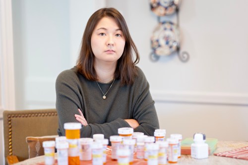 A photo of a woman sitting at a table with several pill bottles in front of her.