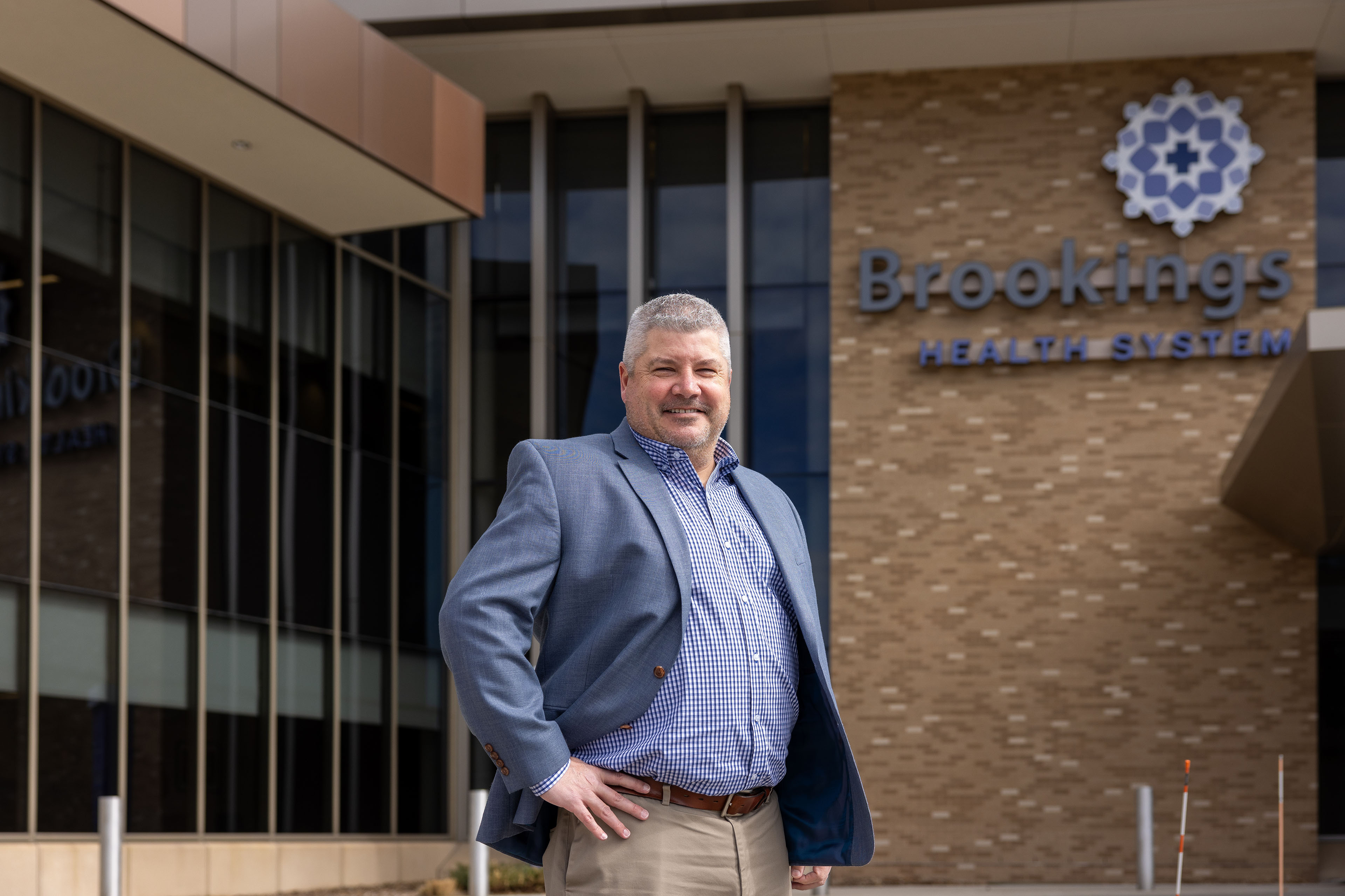 A man in business attire, with short-cut greying hair, stands in front of a hospital building. With his hands on his hips, he smiles. Behind him, building signage reads: "Brookings Health System"