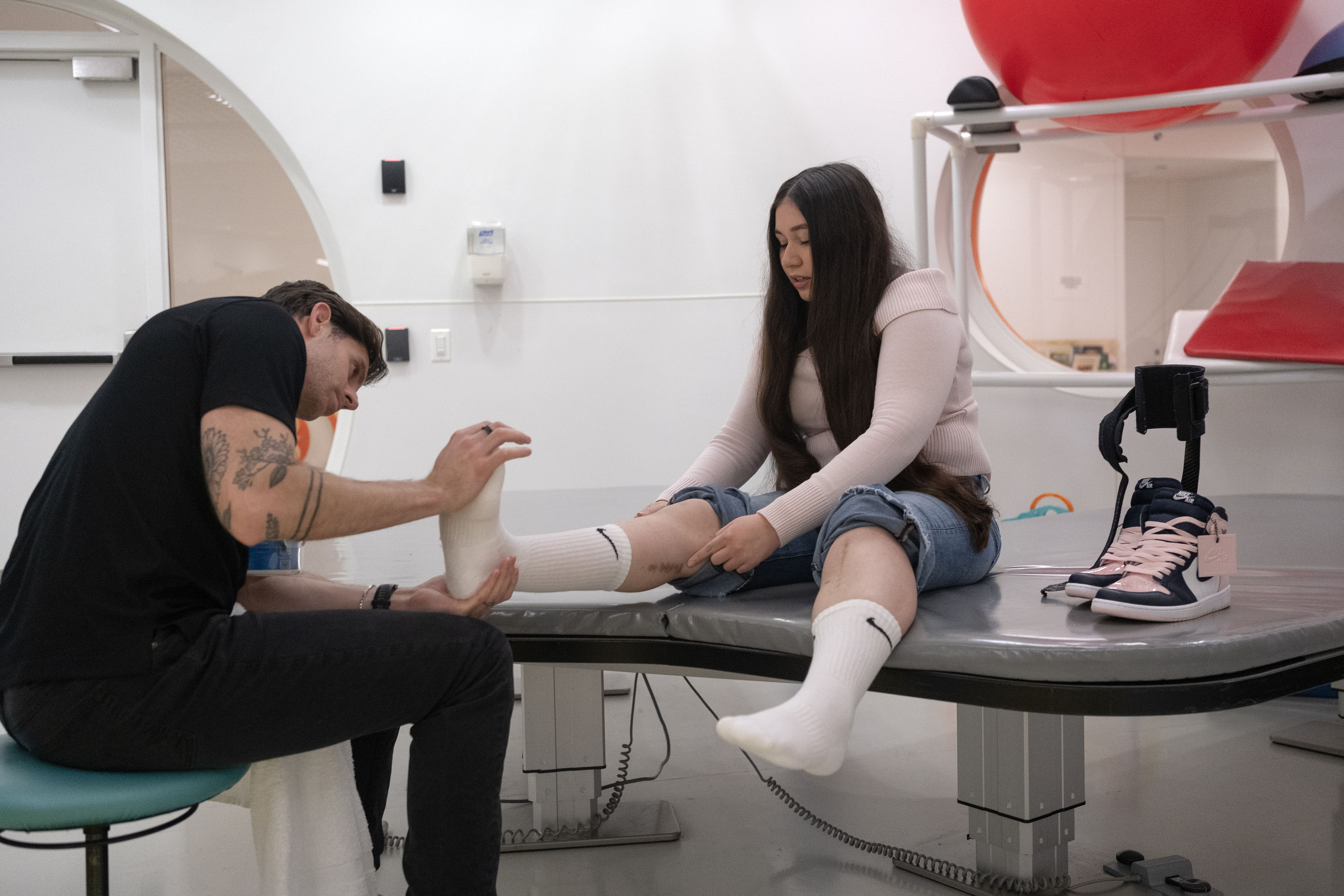 A physical therapist wearing a black shirt and pants presses the foot of a woman sitting on a table
