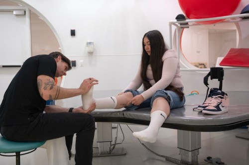 A physical therapist wearing a black shirt and pants presses the foot of a woman sitting on a table