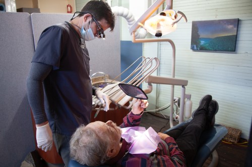 A photo of a dentist working with his patient, who is lying back in a dental chair, using a mirror to look at his teeth.