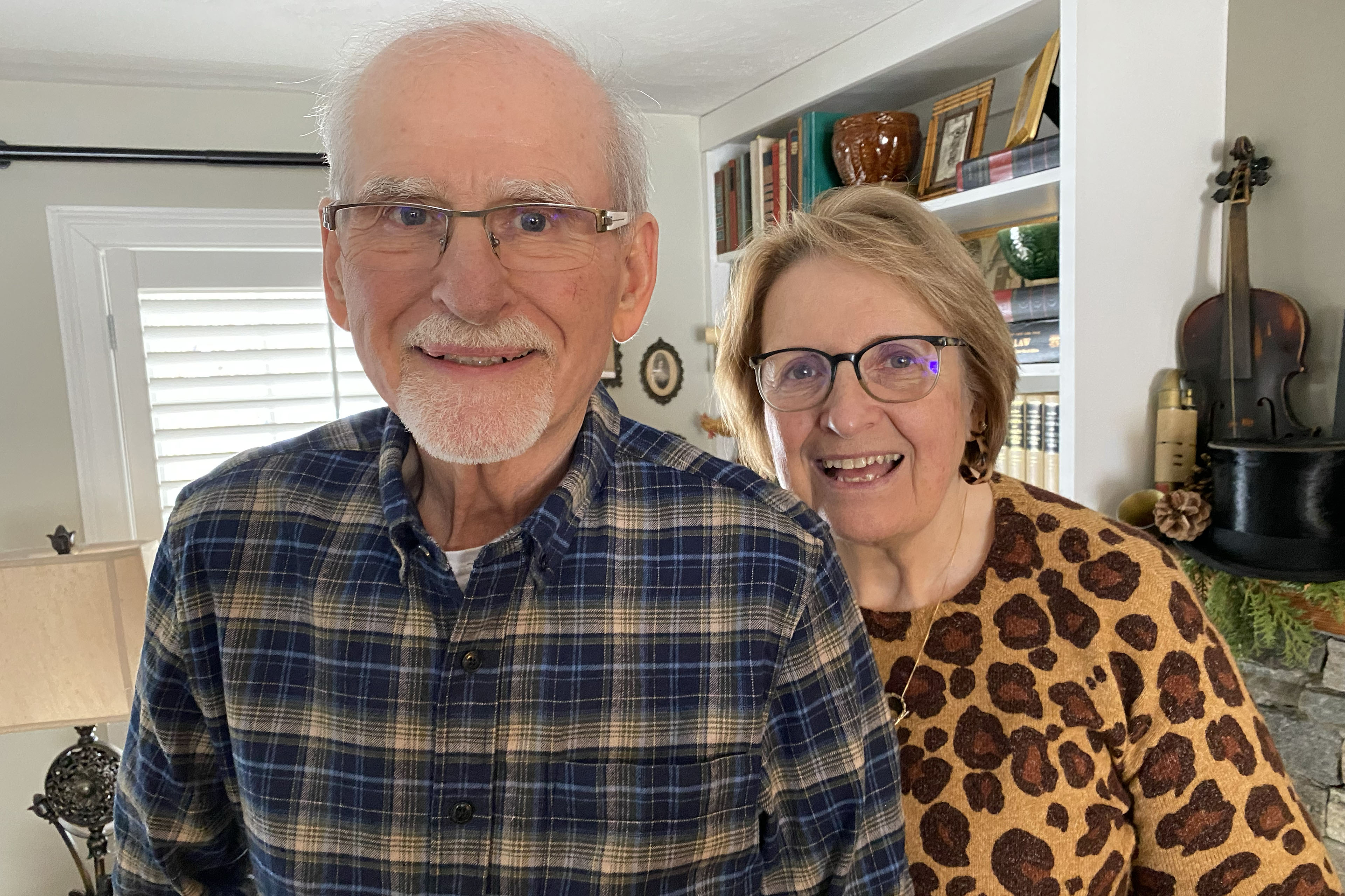 A senior man and woman smile at the camera. They are in their home.