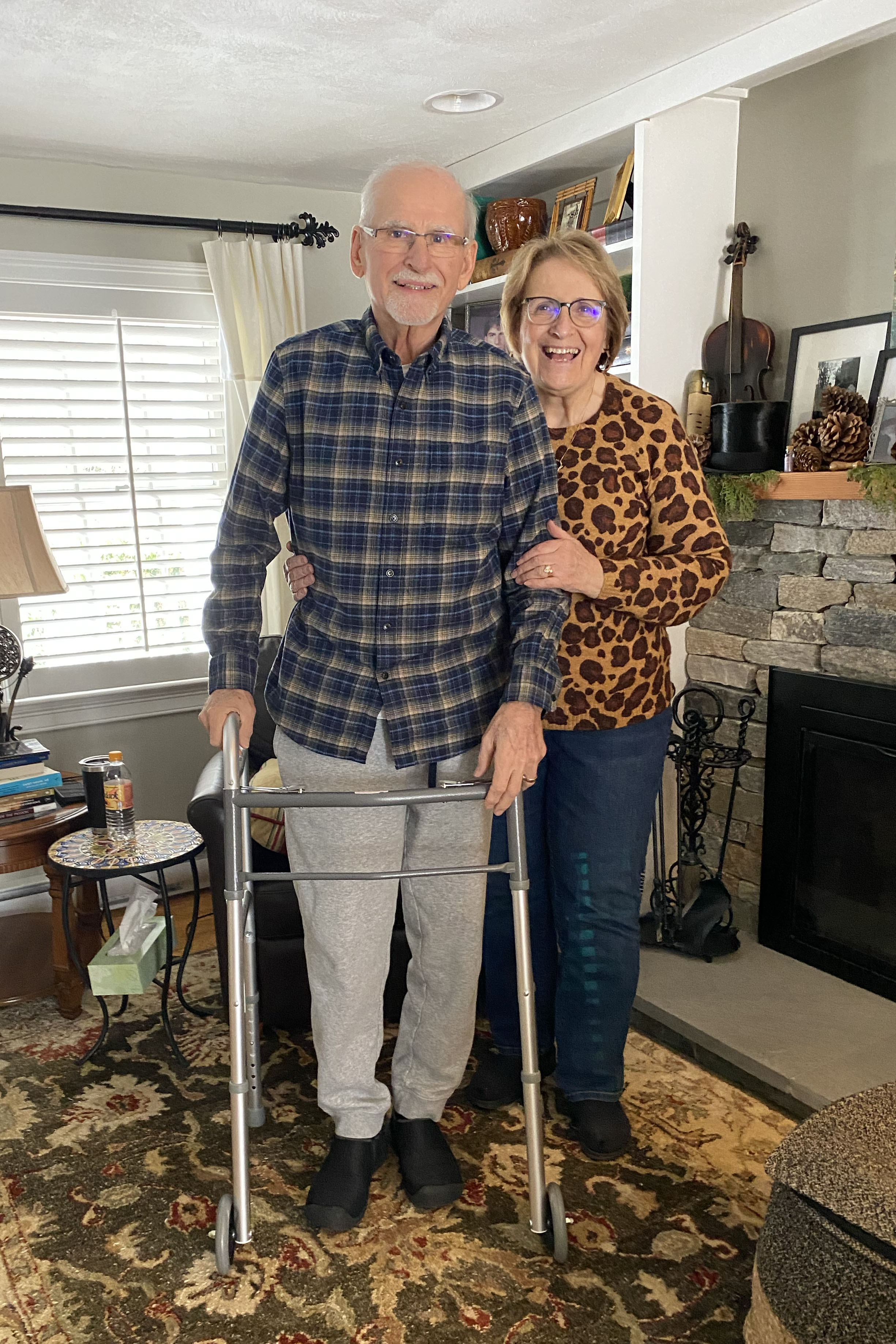 A senior man stands holding a walker. His wife stands beside him and holds his arm and waist. They both are smiling at the camera.