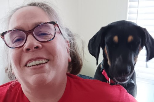 A woman in a red shirt and glasses smiles at the camera as a dog behind her looks over her left shoulder.