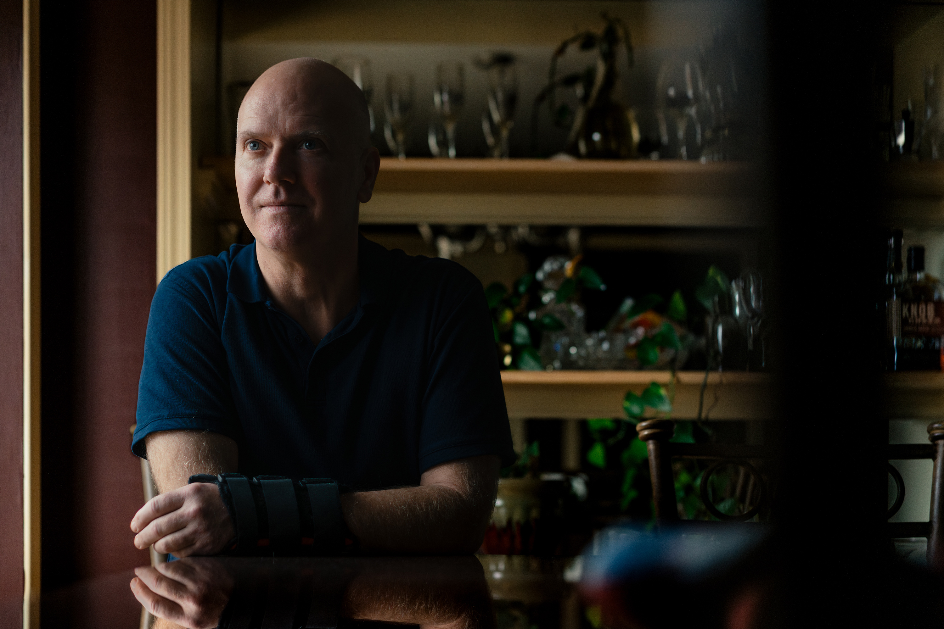 A photo of Tim Winard sitting at a table beside a window. His face and much of his surroundings are cast in shadow.