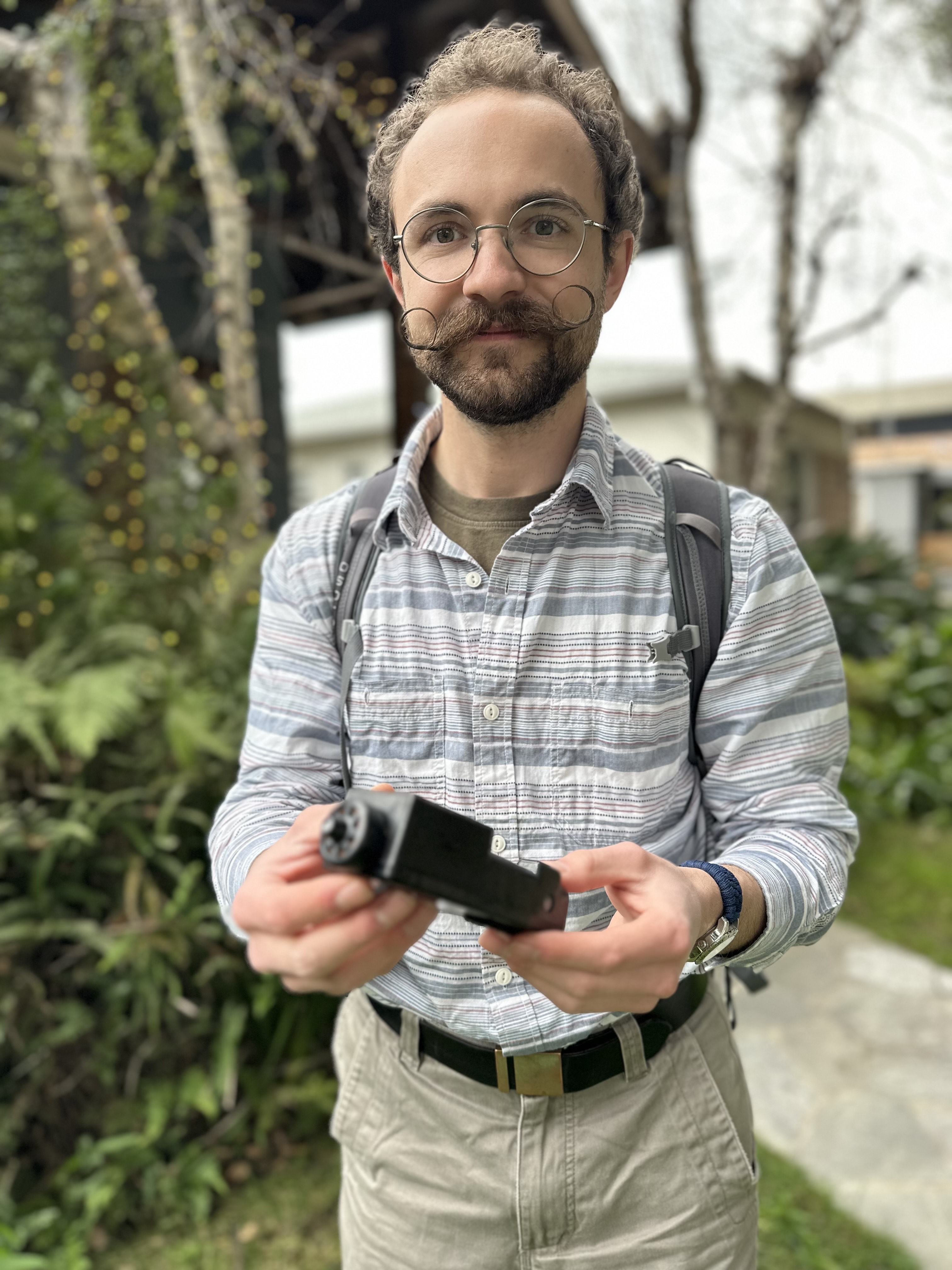 A man stands outside holding a black device and looks at the camera.