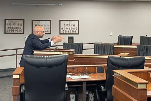 A photo of lawyer Aaron Siri gesturing while speaking at a lectern in a hearing room.